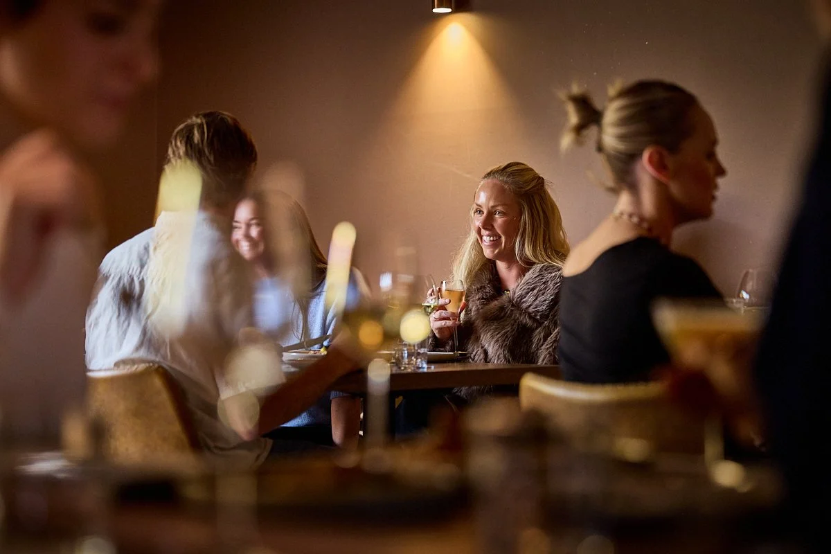 Woman holding a drink sitting at a table in a busy restaurant, smiling at her partner, with blurred outlines of other diners, glasses, and dishes in the foreground