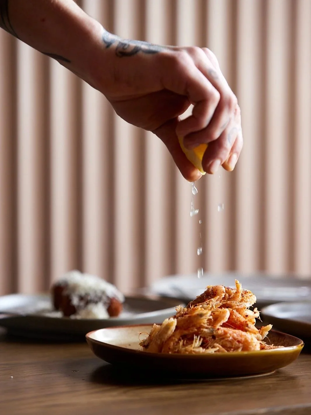 A hand squeezing lemon juice over a plate of fried school prawns, with other plates of food in the background