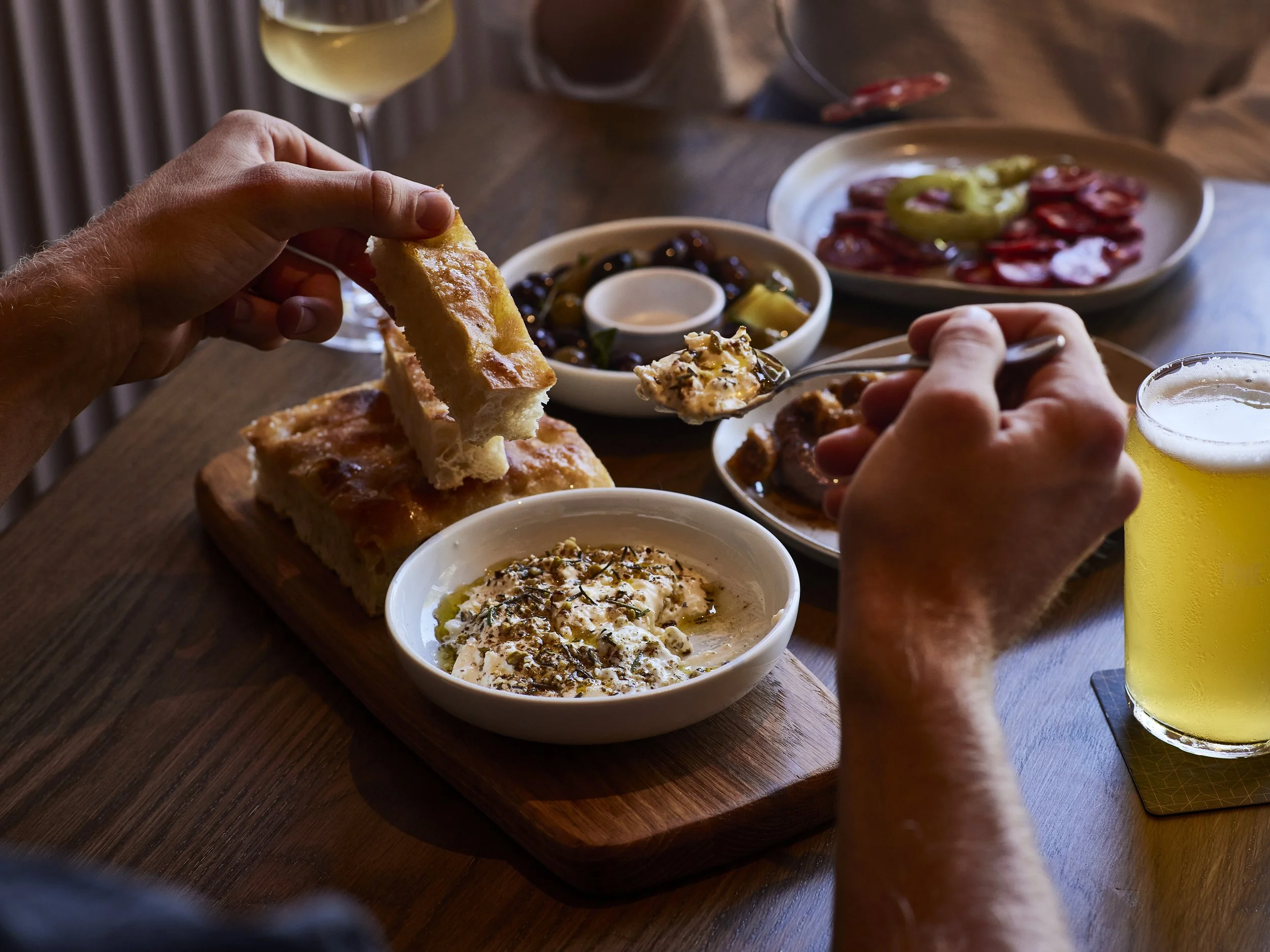 People enjoying a meal with bread, dips, and charcuterie at a wooden table, accompanied by glasses of white wine and beer.