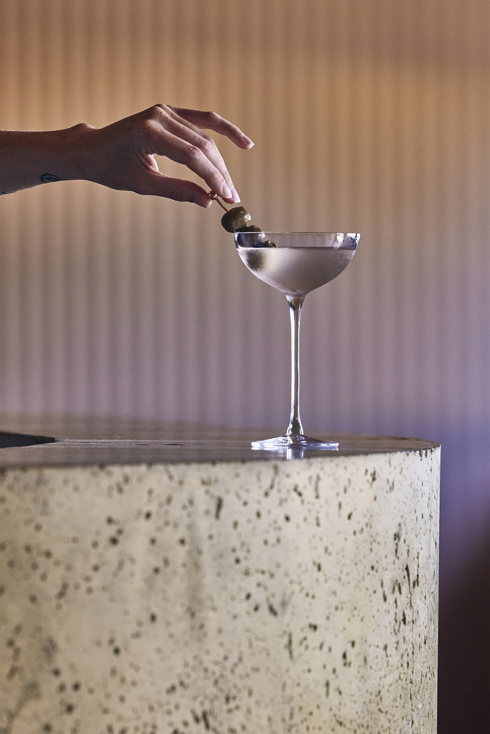 Hand placing three green olives on a skewer into a martini glass with clear liquid, set on a textured beige surface against a gradient background.