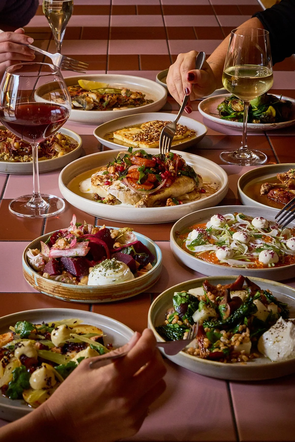 A large selection of dishes laid out on a table, with a couple glasses of wine laid out in between them. A few hands holding forks are reaching in to pick up food