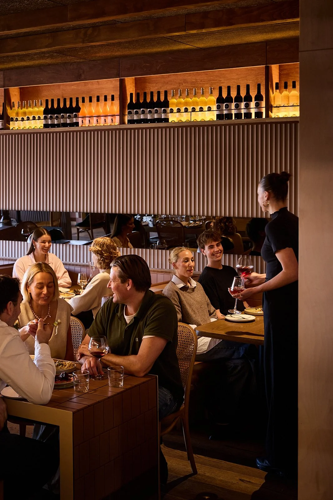 A lively restaurant scene with groups of people dining, wine glasses, and a waiter serving. Warm lighting, wooden accents, and shelves of wine bottles add to the cozy ambiance.