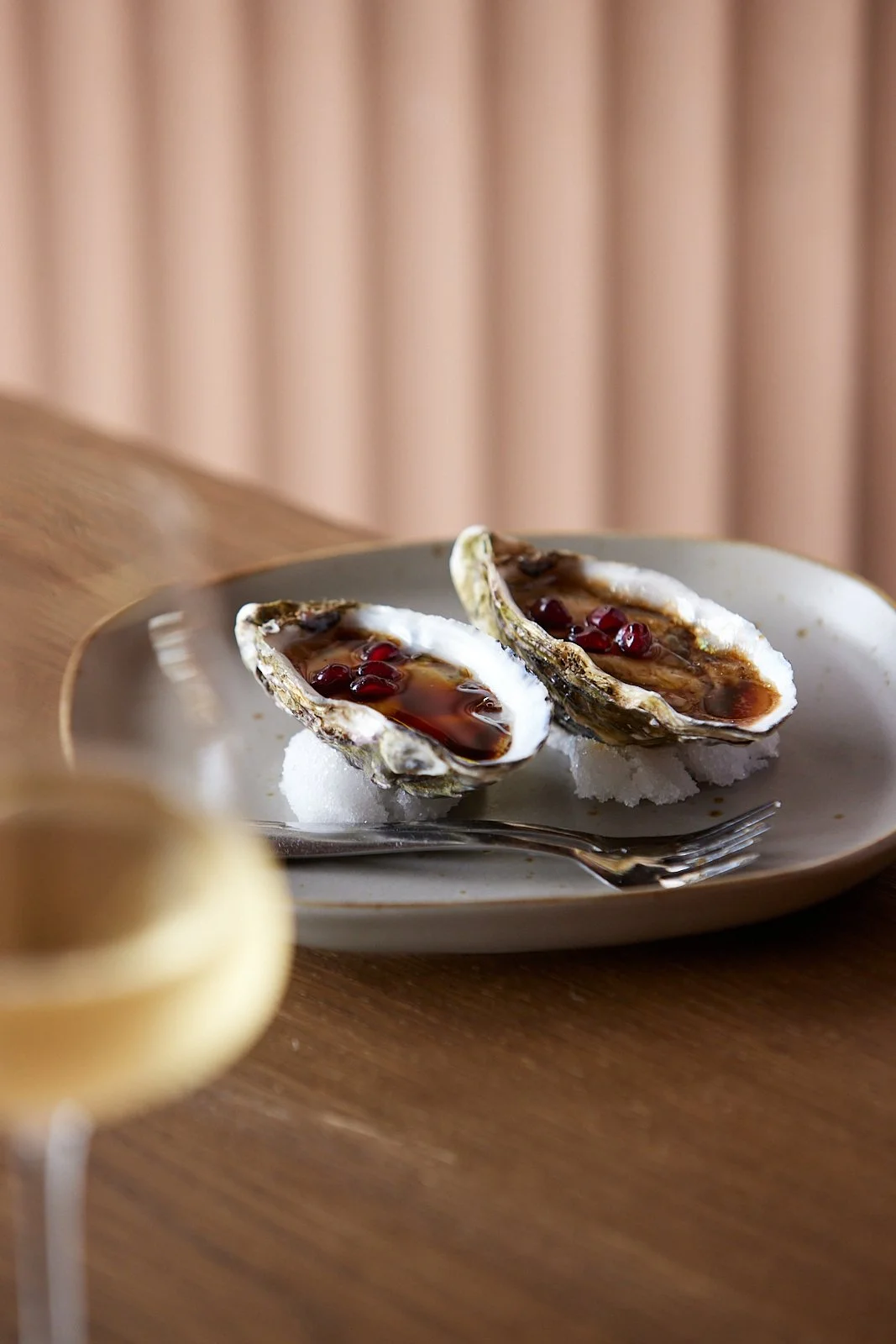Two oysters on the half shell topped with a dark sauce and pomegranate seeds, served on a white plate with a fork, on a wooden table