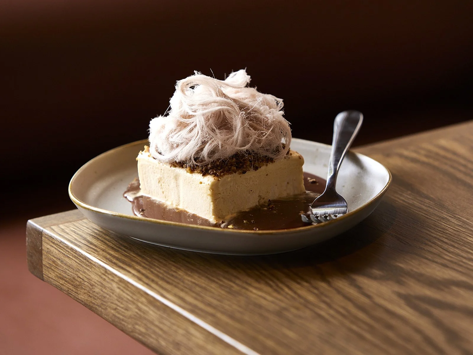 Peanut butter parfait topped with baklava spiced nut crumb, pashmak (a persian fairy floss), and sitting on salted caramel on a plate on a wooden table.