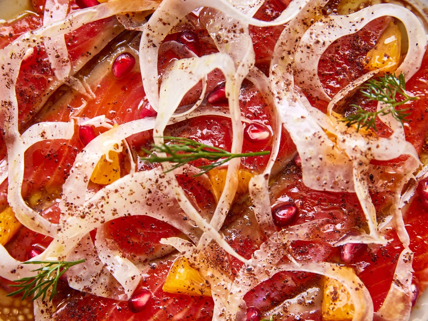 Close-up of a cured fish dish, with fennel ribbons, pomegranate seeds, orange cubes, and garnished with herbs