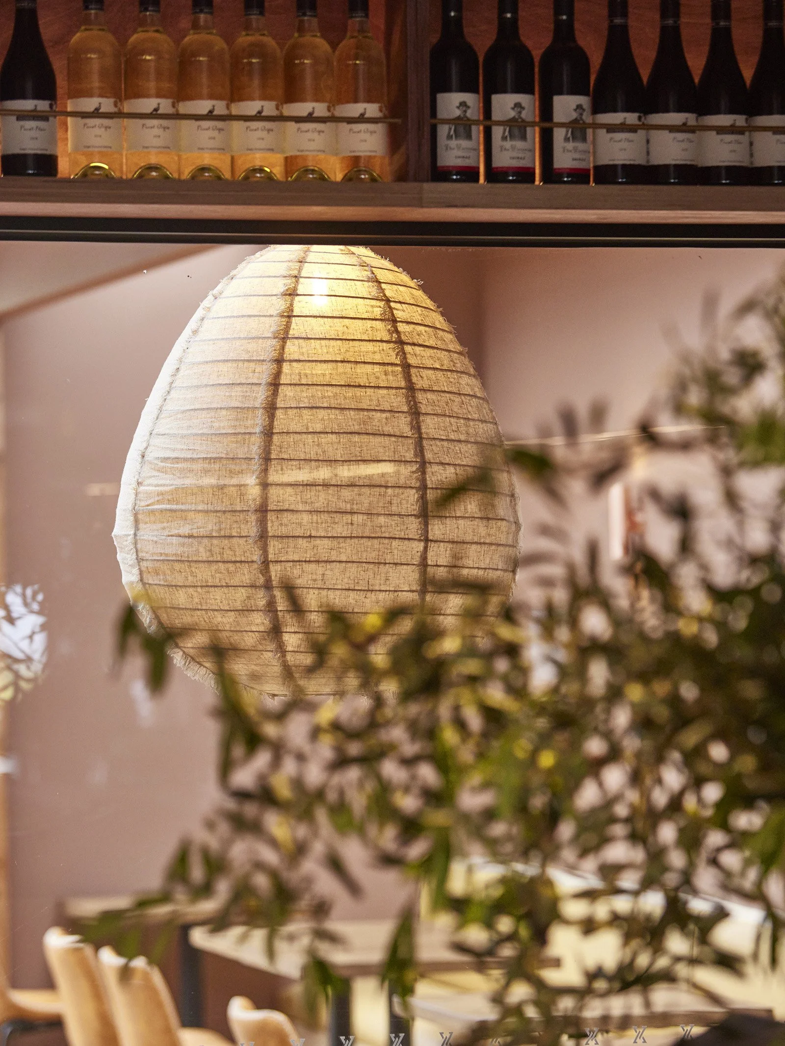 A warm-colored fabric paper lantern hanging from the ceiling in an indoor space, with blurred flowers and chairs in the background.