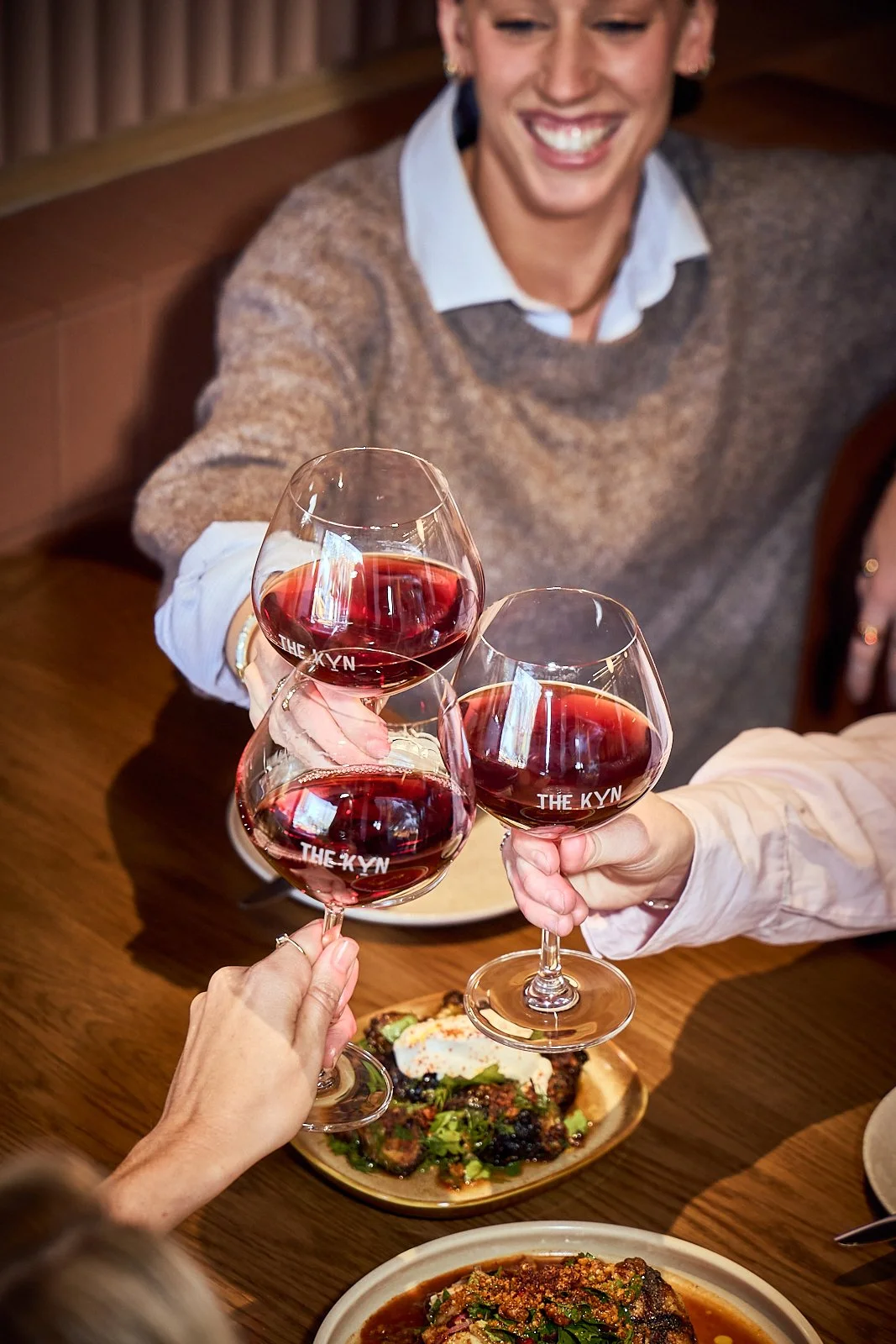 People clinking glasses of red wine during a meal at a restaurant, with plates of food on the table and a smiling woman in the background.