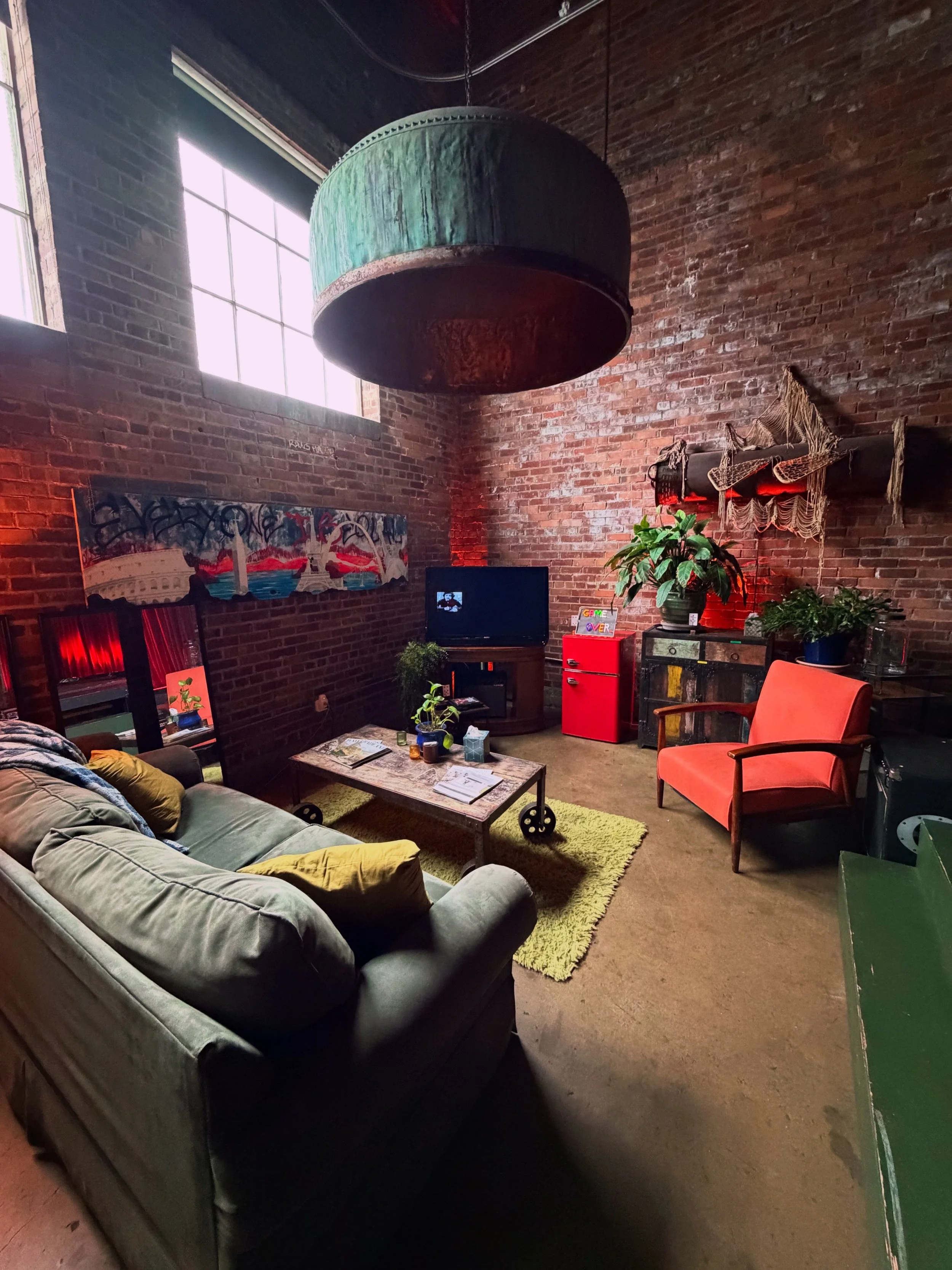 corner of the gastropub with a lounge chair and couch, with a T.V, video games, speaker, bar cart, mini fridge and art.