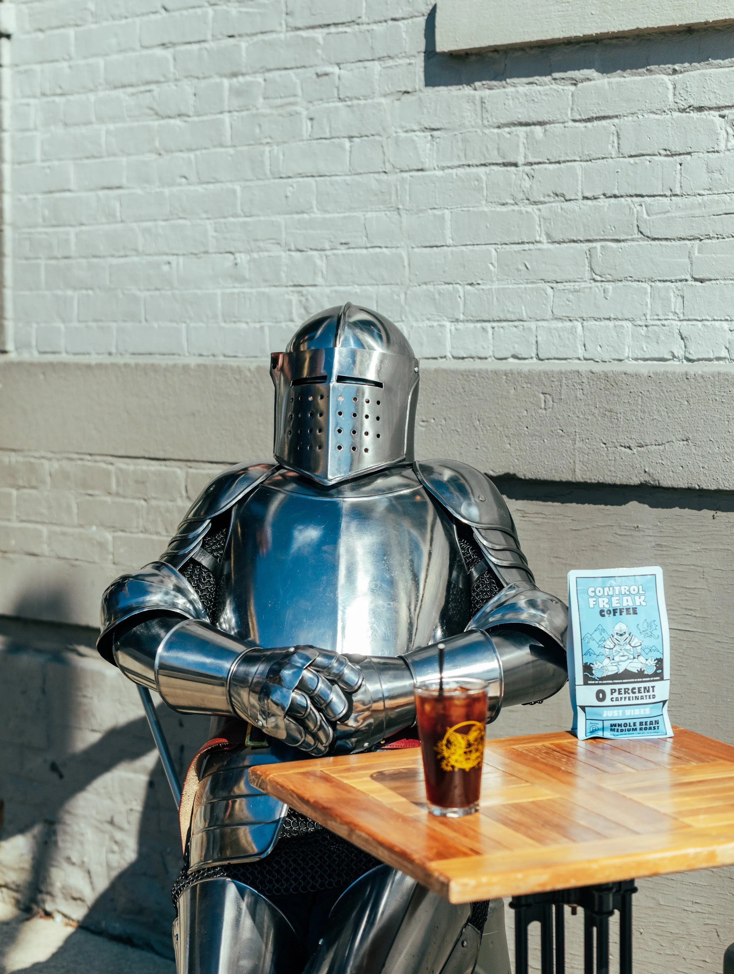 A person dressed in shiny metallic armor, resembling a knight, is sitting at a wooden table with a glass of cold brew in front of them. The armor covers their entire body and head.