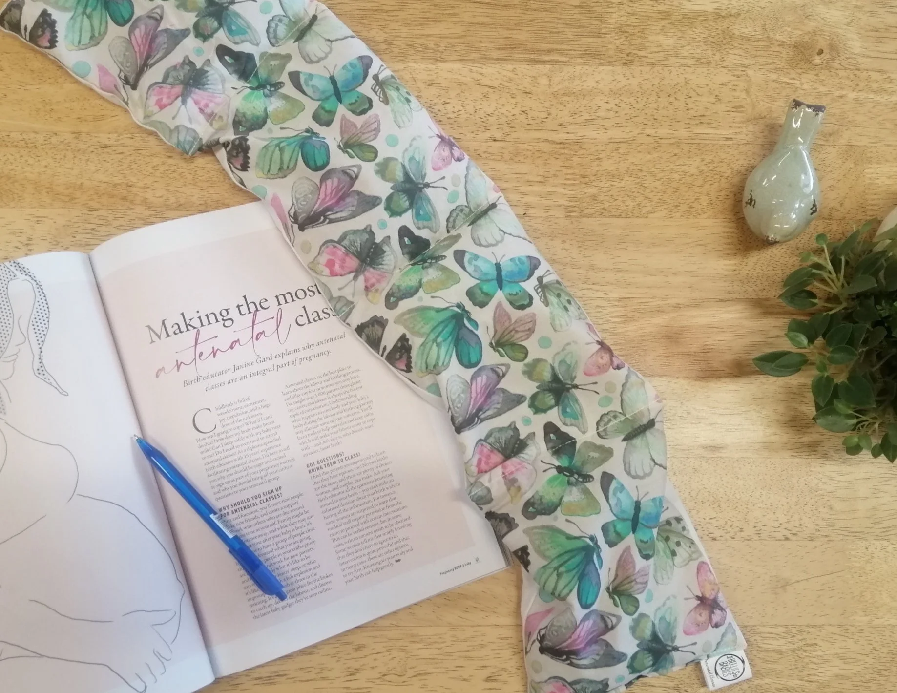 Open magazine with article titled 'Making the most of antenatal classes' on a wooden table, with a butterfly-printed pregnancy wheat bag partially covering the magazine, a blue pen on the page, a small decorative bottle, and a potted plant.