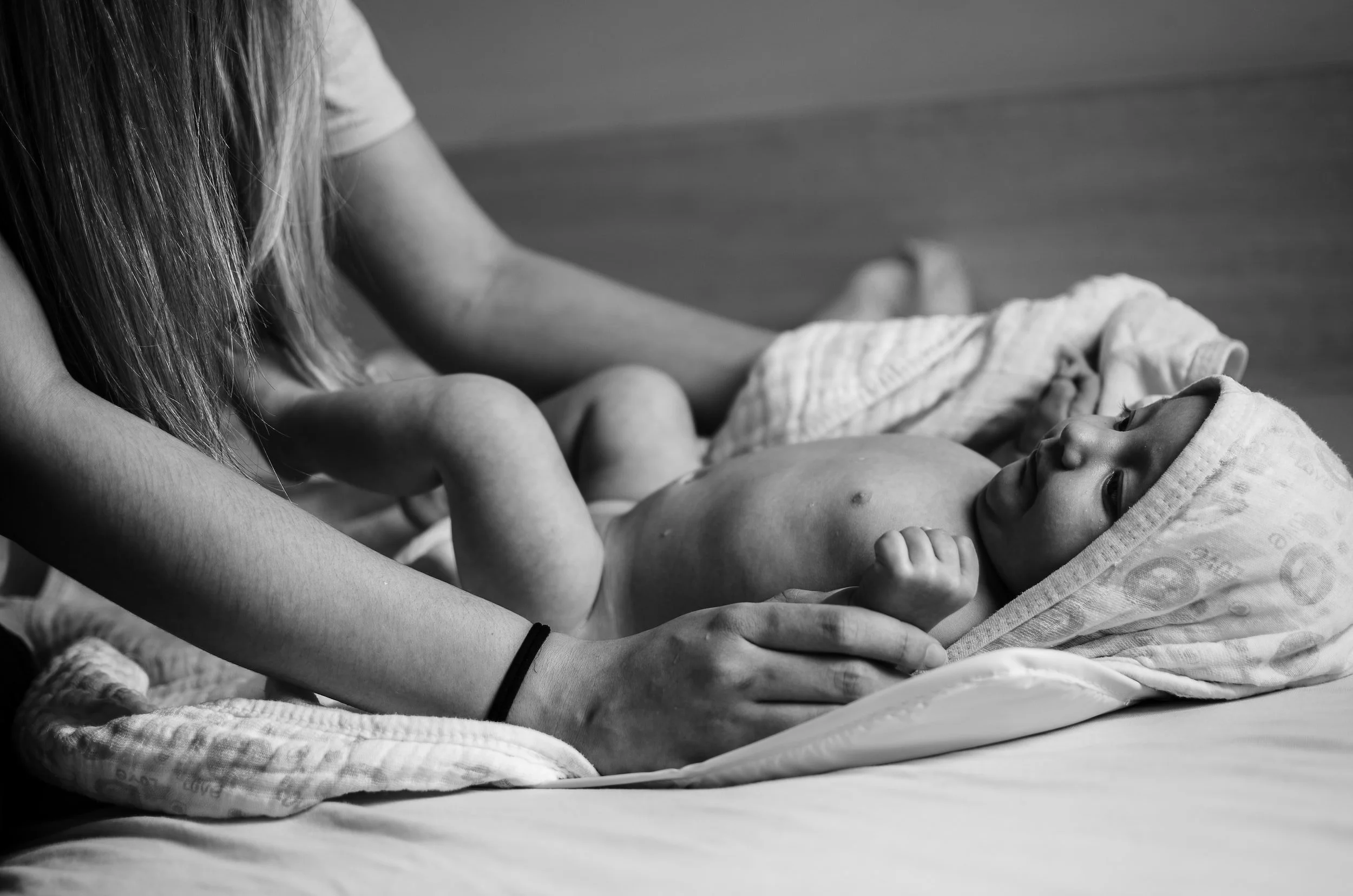 A person laying on a bed, holding a baby who is lying on their back with a towel over their head, both in black and white.