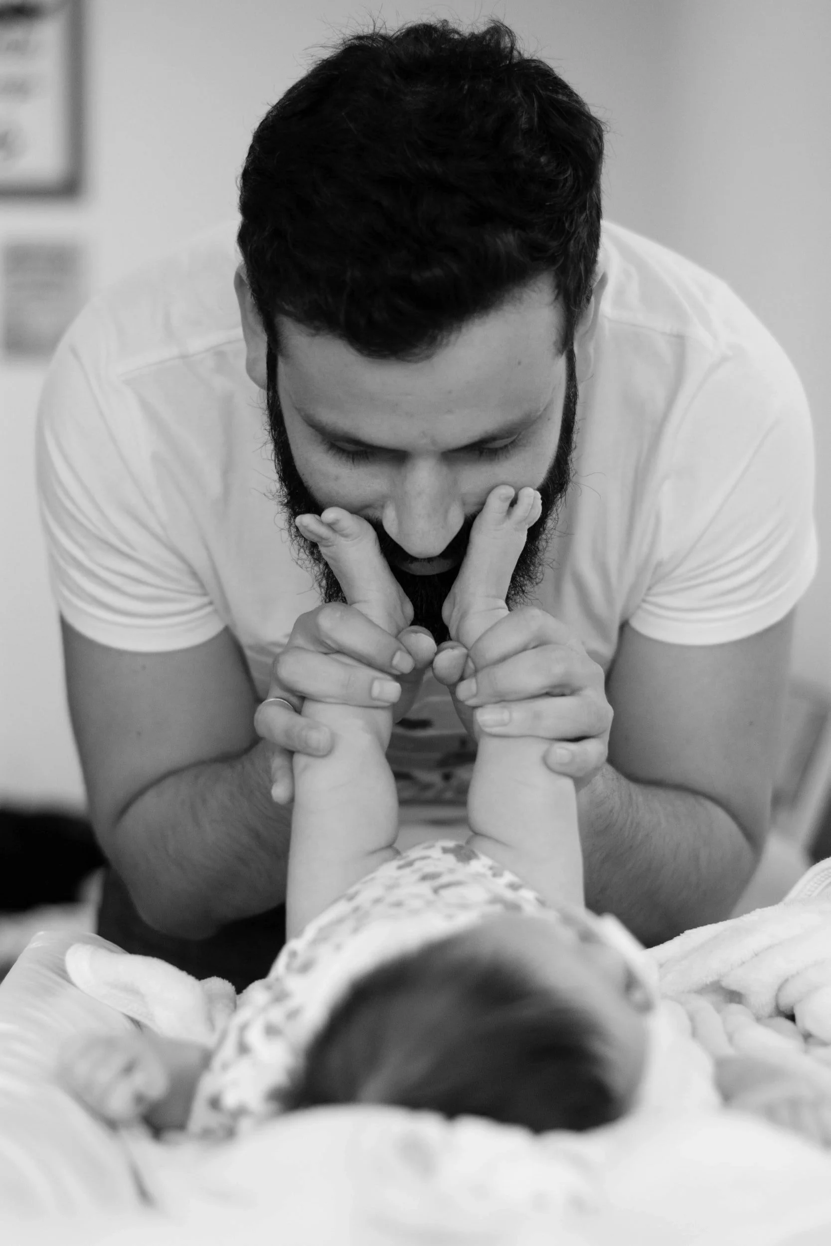 A man with a beard smiling and holding a baby's hands while lying in a hospital bed, with the baby’s face out of focus in the foreground.