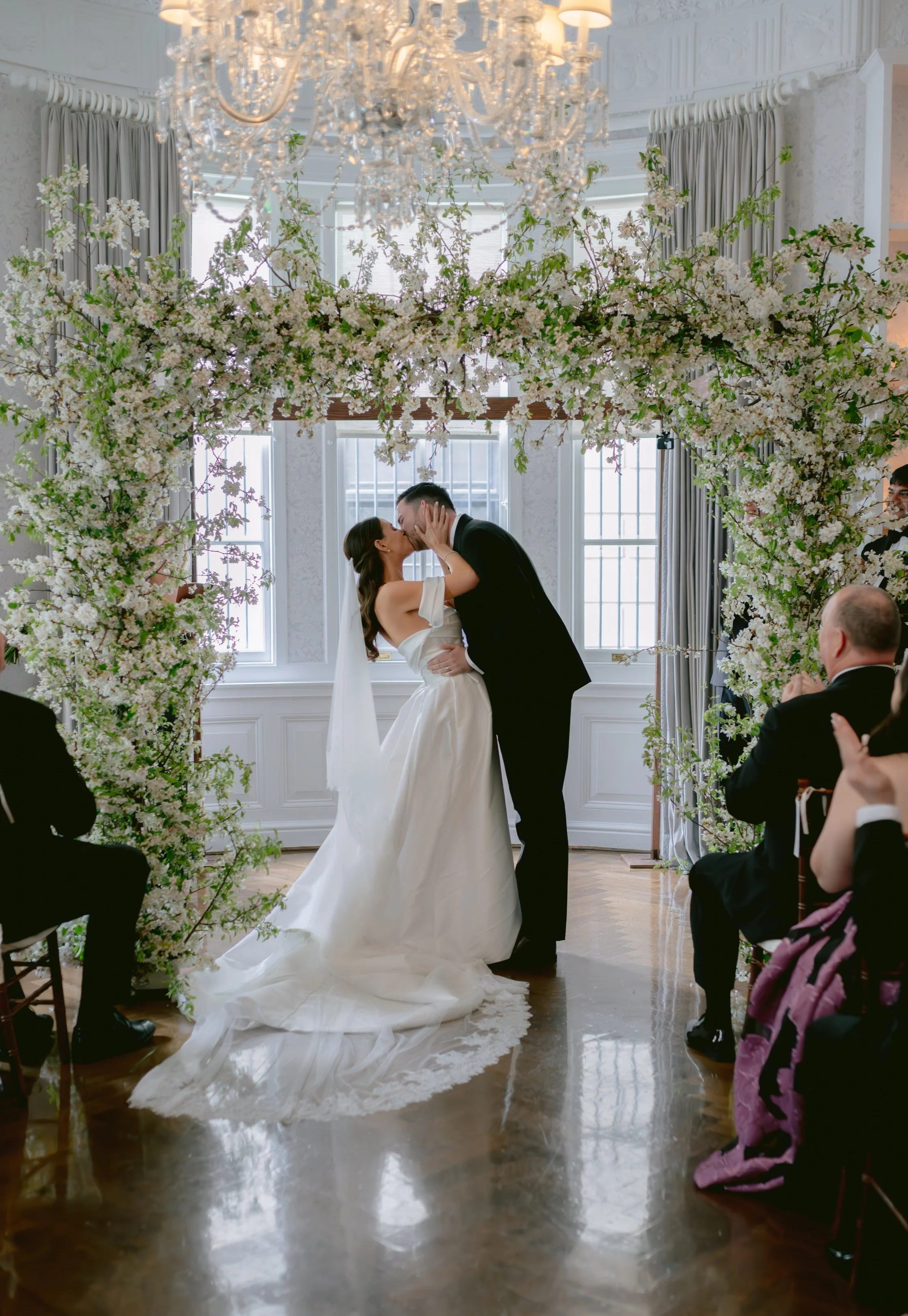 Bride and groom kissing under a floral arch at an indoor wedding ceremony, with guests seated and clapping.
