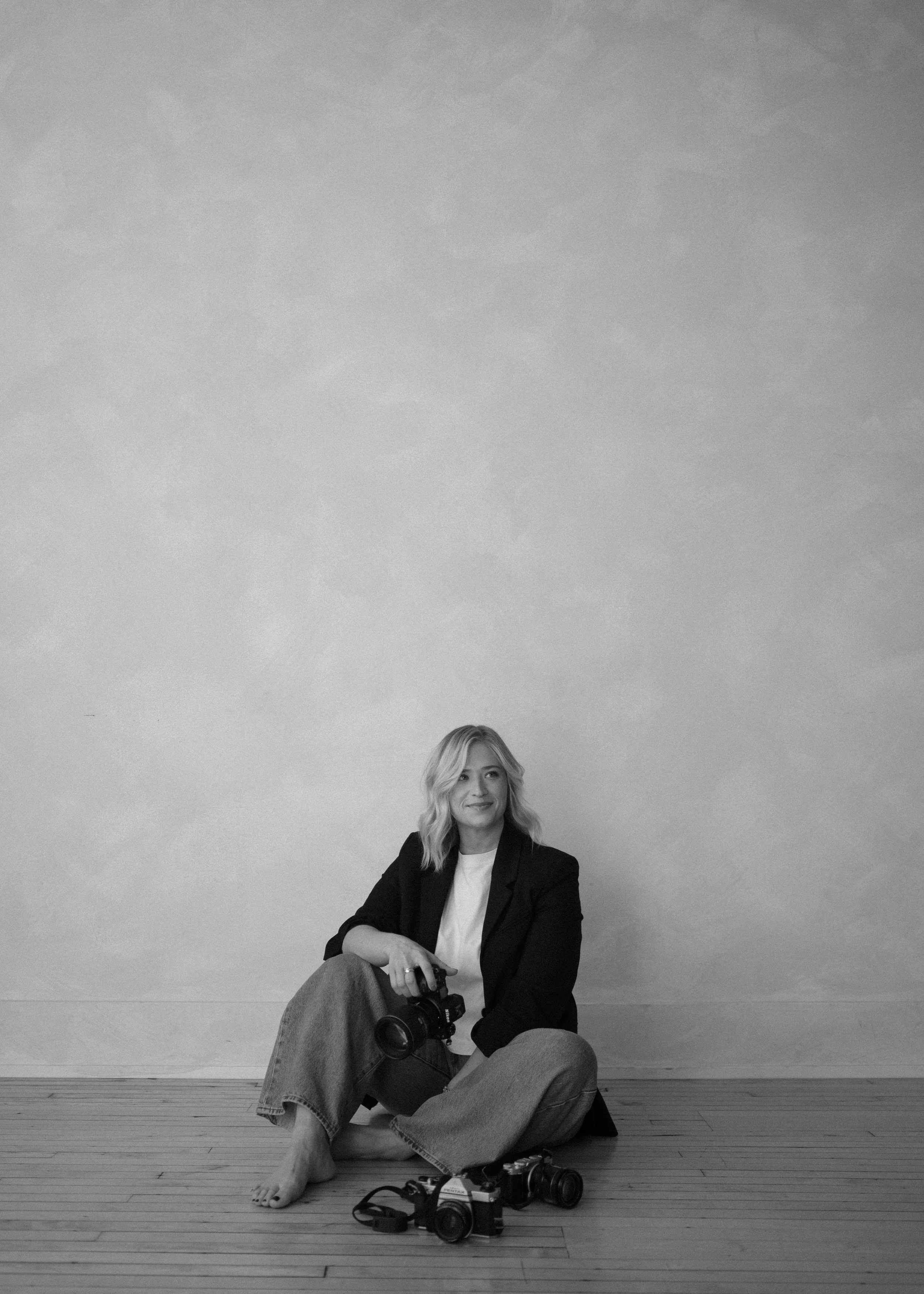 Editorial portrait of Chicago wedding photographer Chandler Austin seated in bright, minimalist studio holding a digital camera with film cameras at her feet, creating timeless, documentary wedding photography in Chicago and destination weddings
