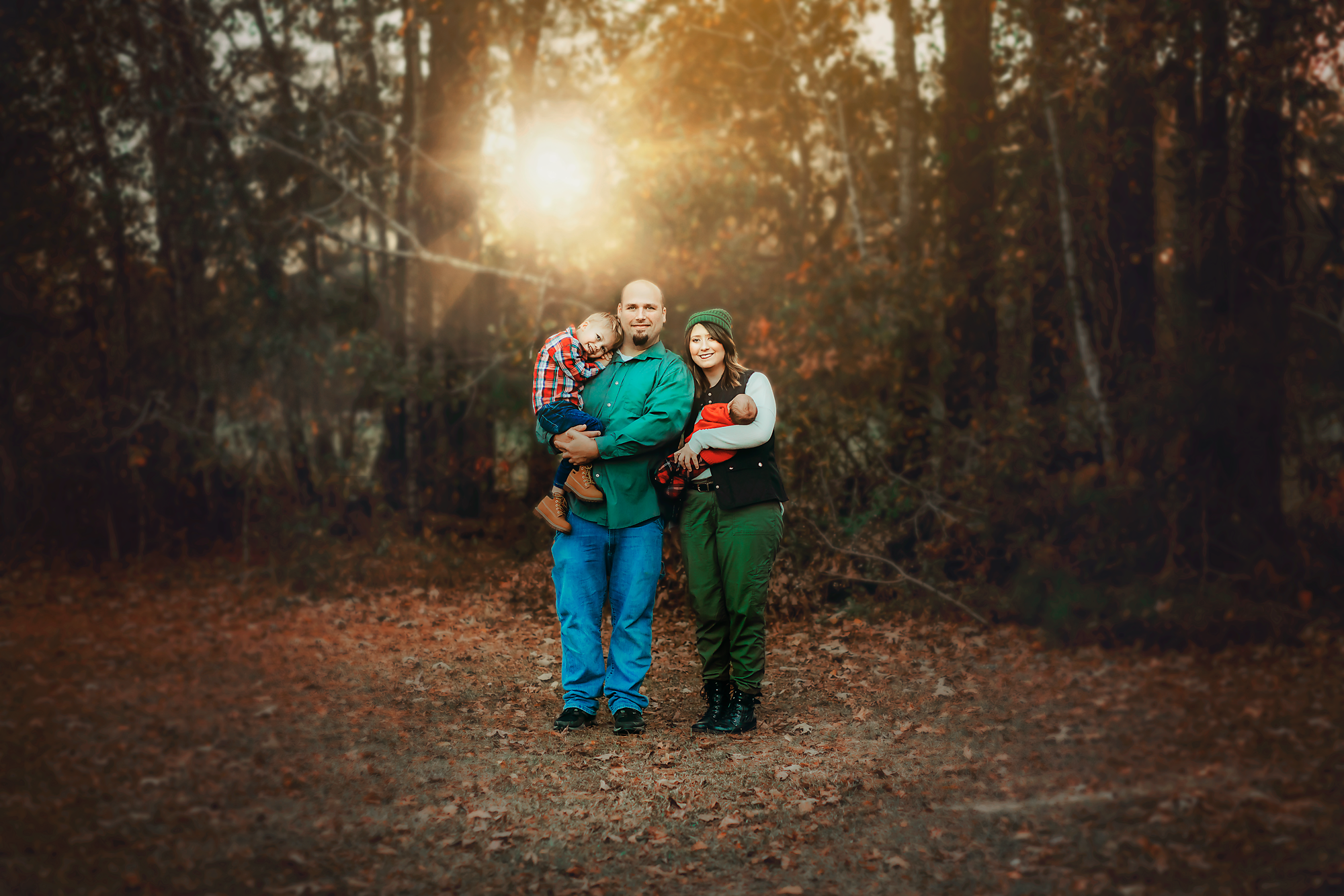 Family of four standing on a wooded trail during autumn, with sunlight filtering through trees behind them. The father is holding a young boy, and the mother is holding a newborn baby.