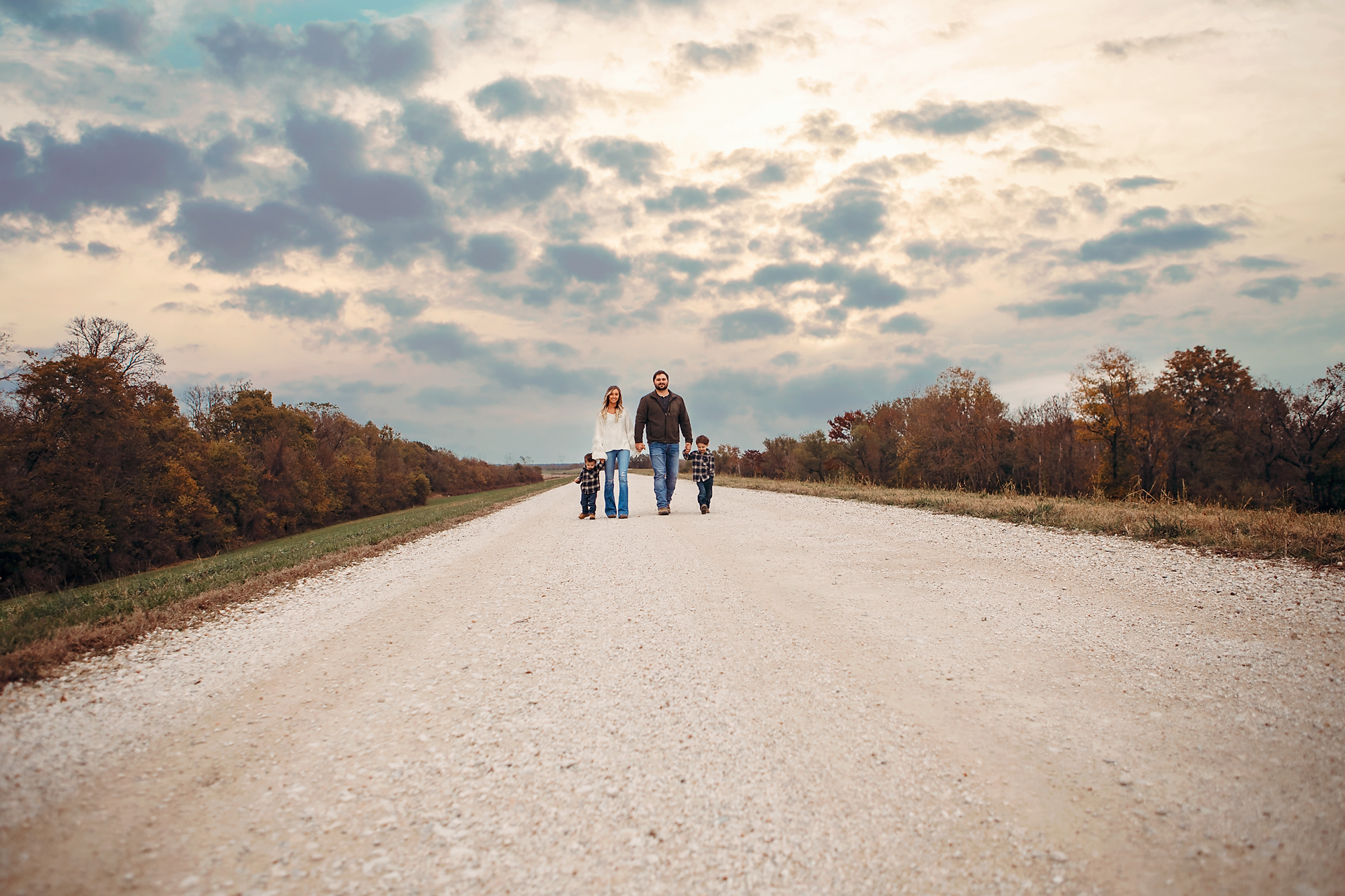 A family of four walking on a wide gravel road in a rural area during autumn, with trees showing fall colors and a cloudy sky overhead.