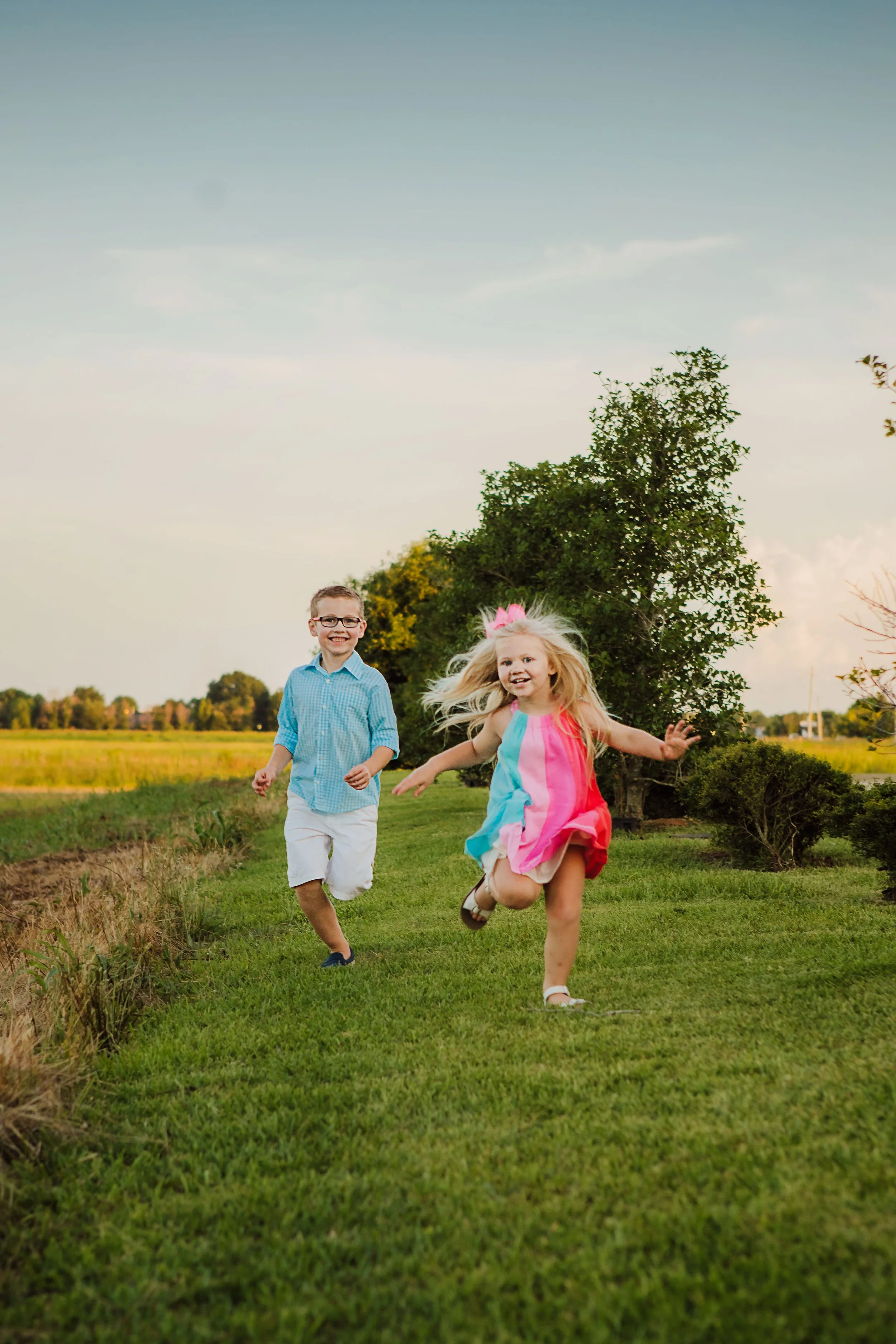 Two children, a boy and a girl, running happily on a grassy field near trees in a rural area on a clear day.