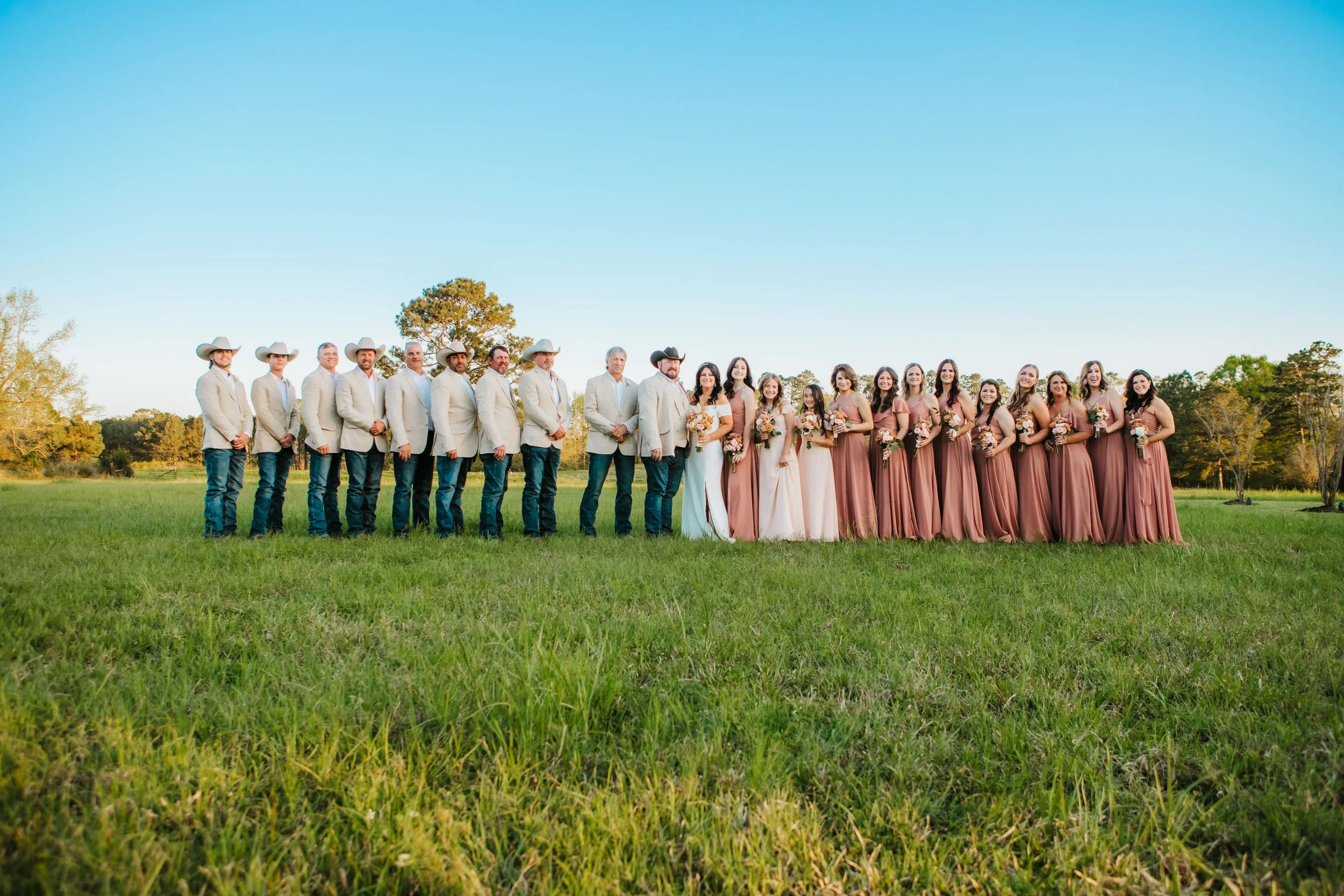 A group of men and women dressed in formal attire standing on a grassy field under a clear blue sky, likely at a wedding or special event.
