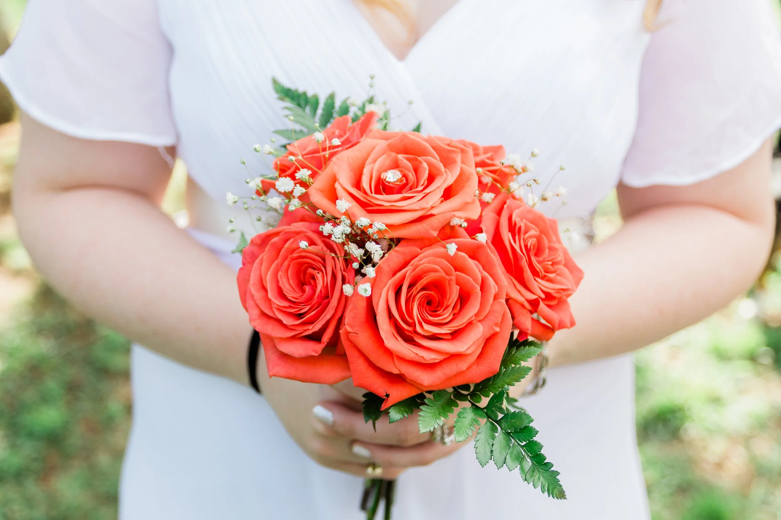 Person holding a bouquet of five orange roses with white baby's breath flowers and green leaves outside.