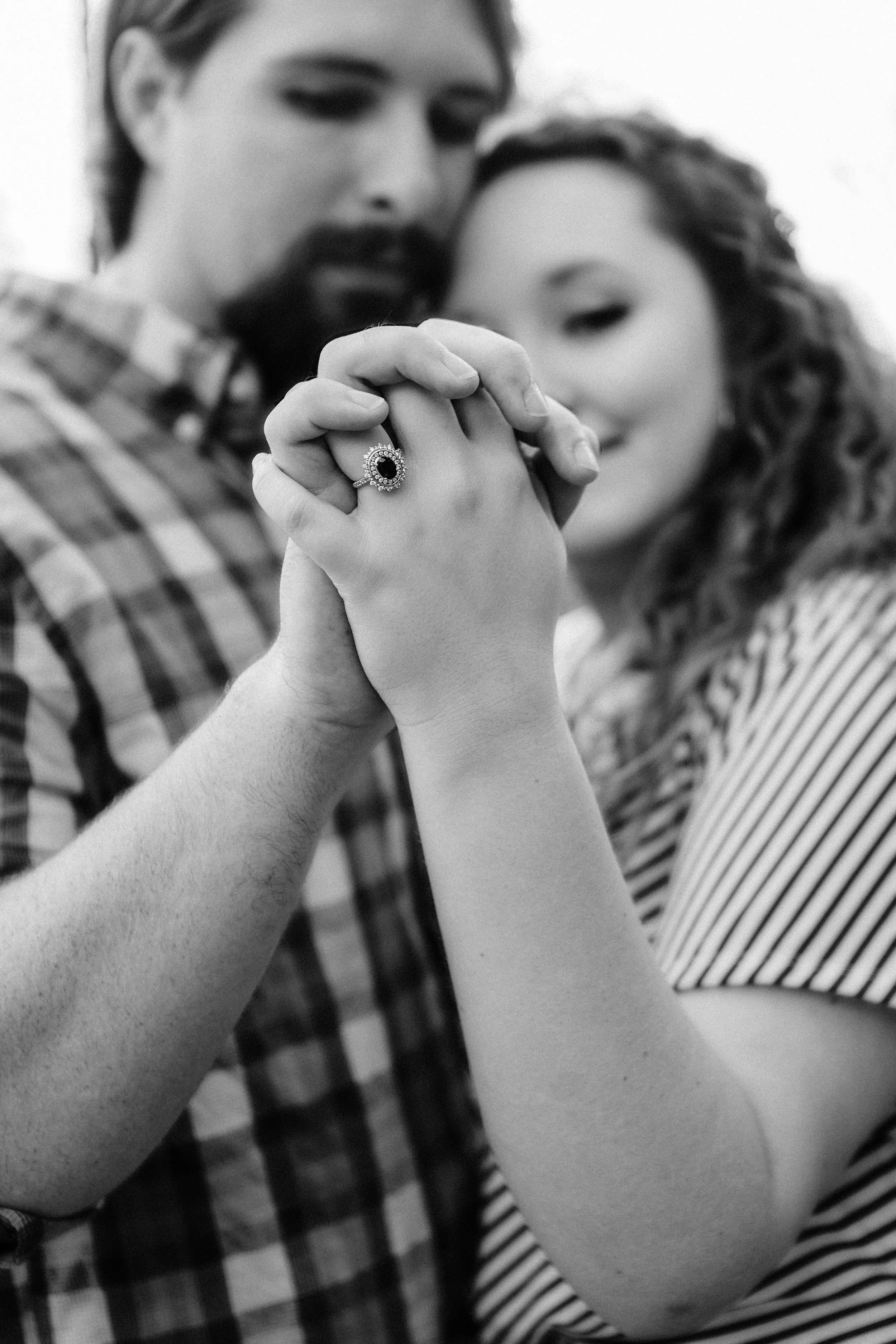 Two people hold hands close together, with a woman wearing an engagement ring, and blurred faces of a man and woman in the background.