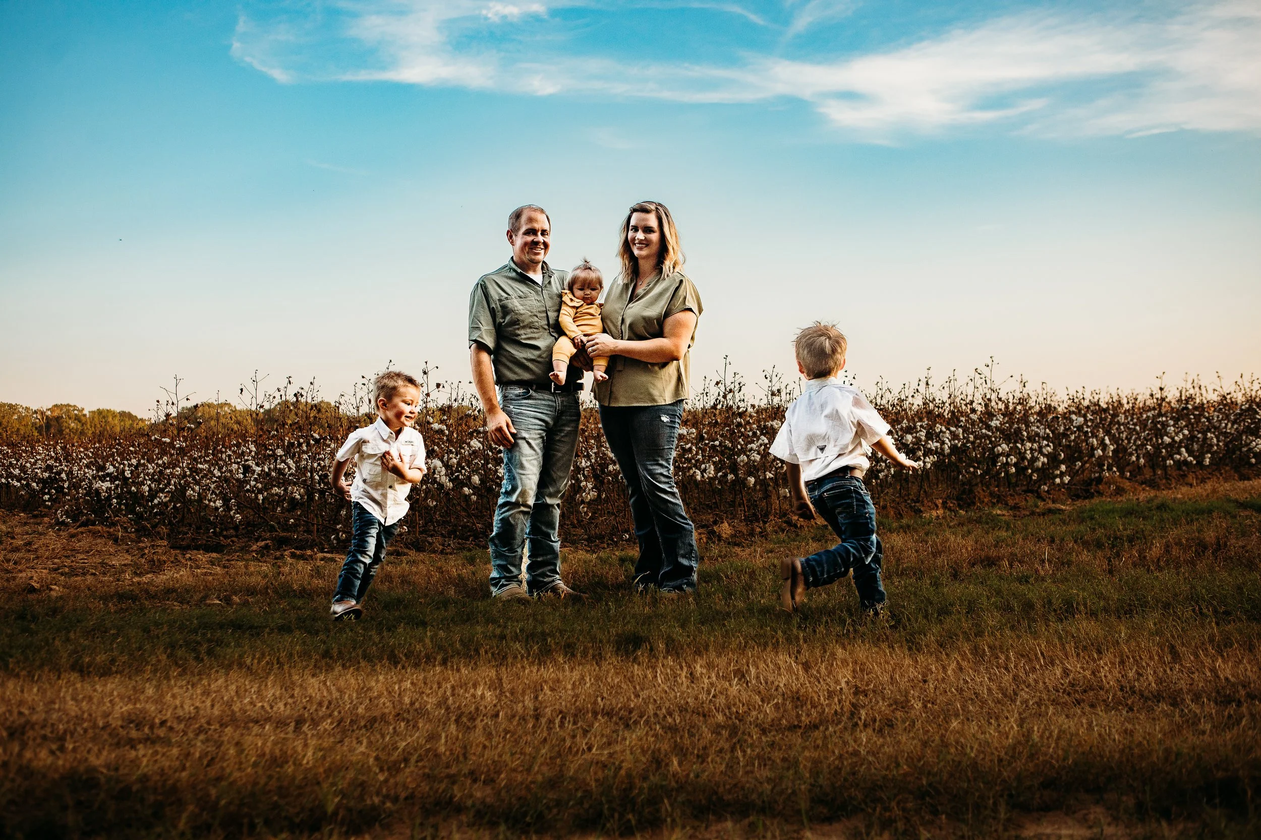 A family of five standing and playing outdoors in a cotton field at sunset, with a clear sky and trees in the background.