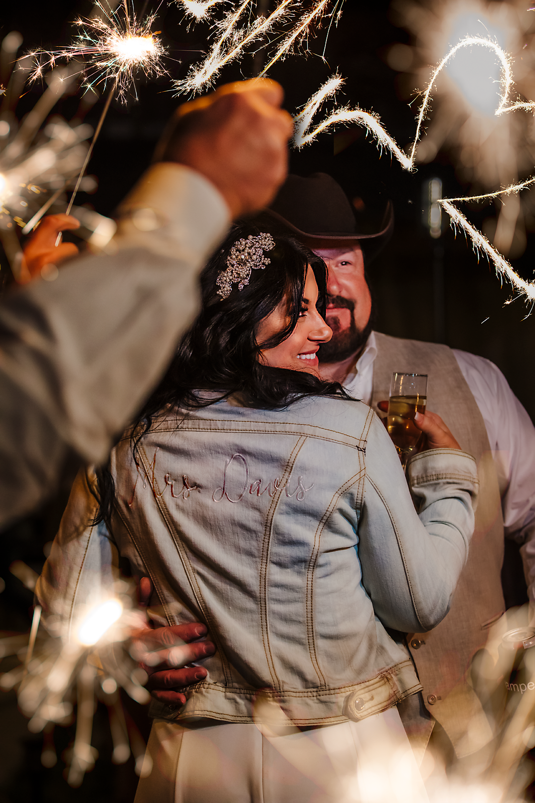 A couple celebrating with sparklers, holding drinks, smiling warmly, with the woman wearing a jacket embroidered with 'Mrs. Davis' and a woman in the background wearing a western hat.