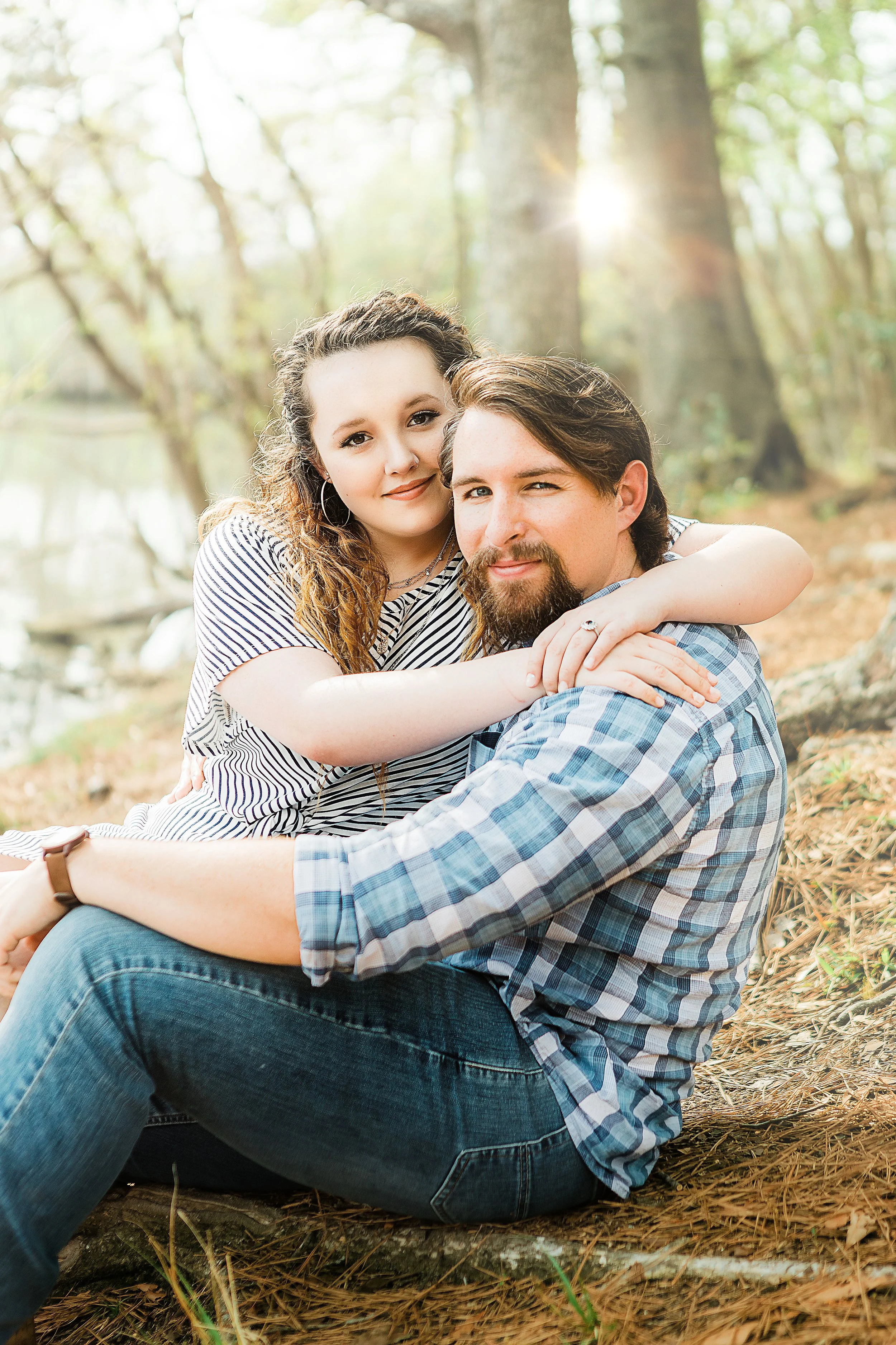 A young woman and man sitting on the ground in a wooded area, with sunlight filtering through the trees, embracing and looking at the camera.