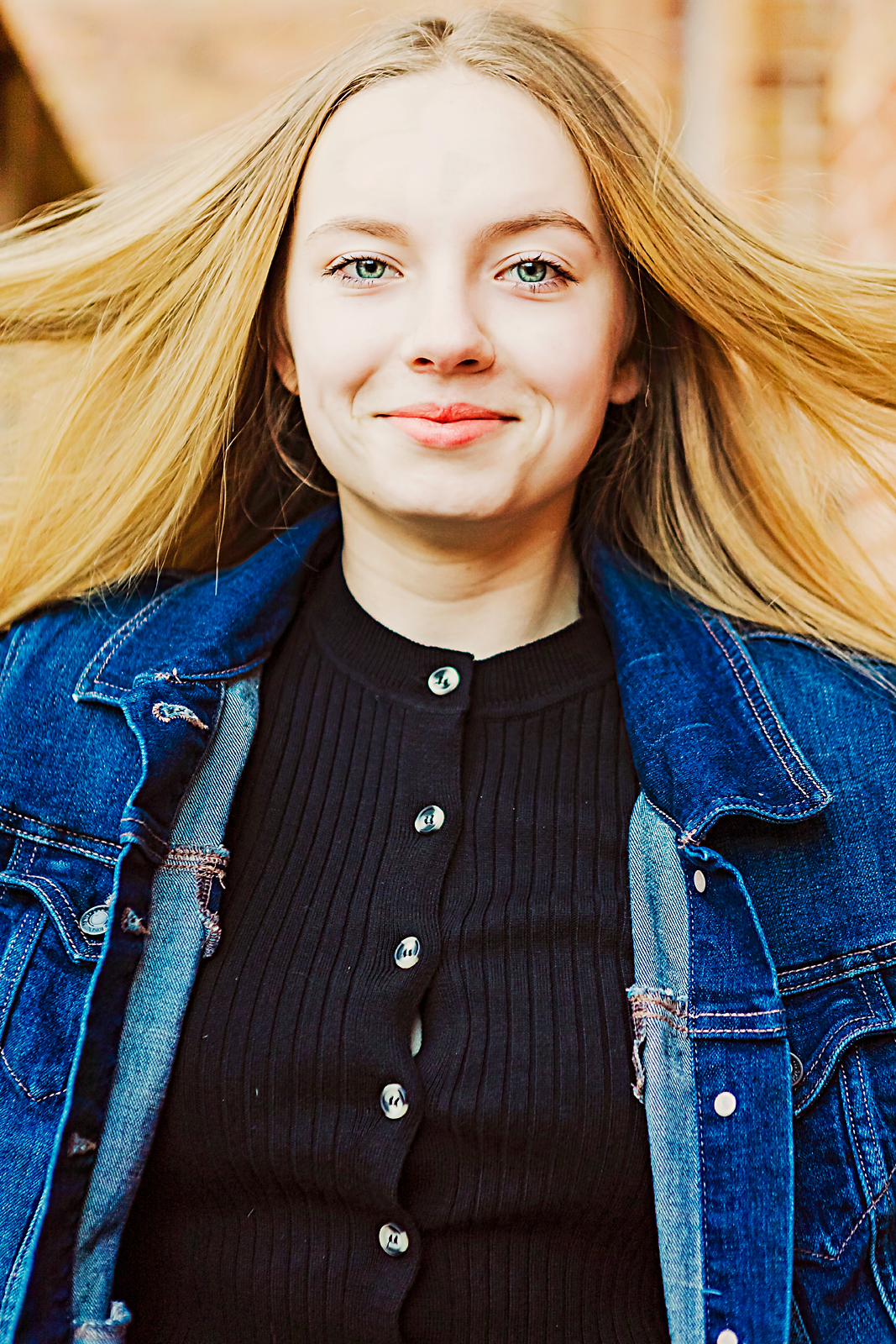 A young woman with long blonde hair and blue eyes smiling, wearing a black ribbed shirt and a blue denim jacket, outdoors.