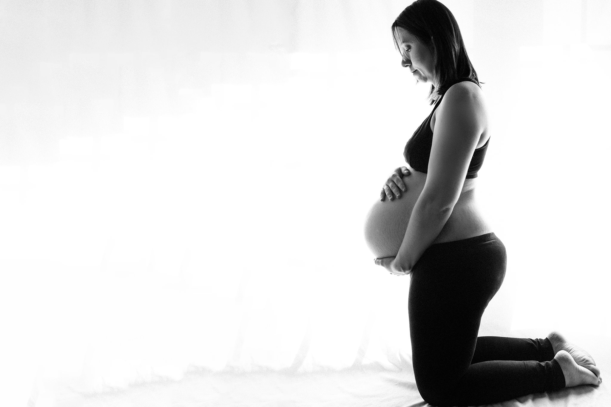 A pregnant woman kneeling on the floor, holding her belly with both hands, looking down, in a black and white photo.
