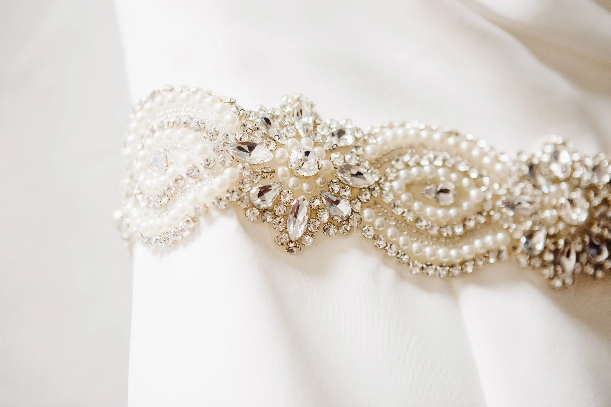 Close-up of a pearl and crystal embellishment on a white satin fabric, likely part of a bridal accessory or gown embellishment.