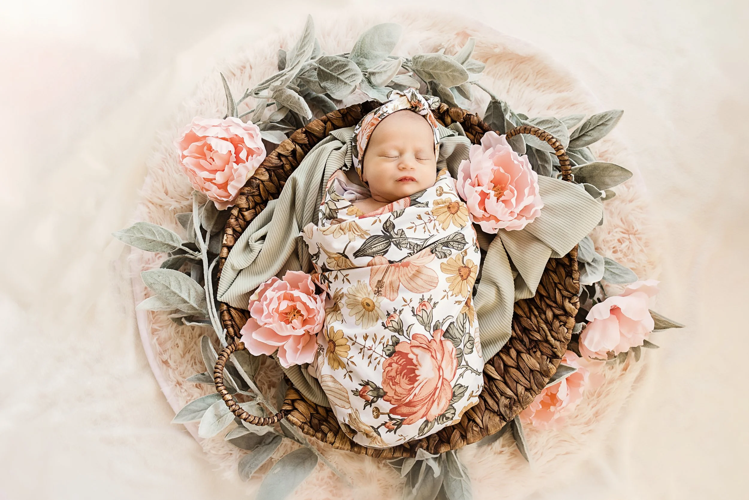 A newborn baby sleeping in a basket, wrapped in a floral blanket, surrounded by pink flowers and green leaves on a soft pink fuzzy blanket.