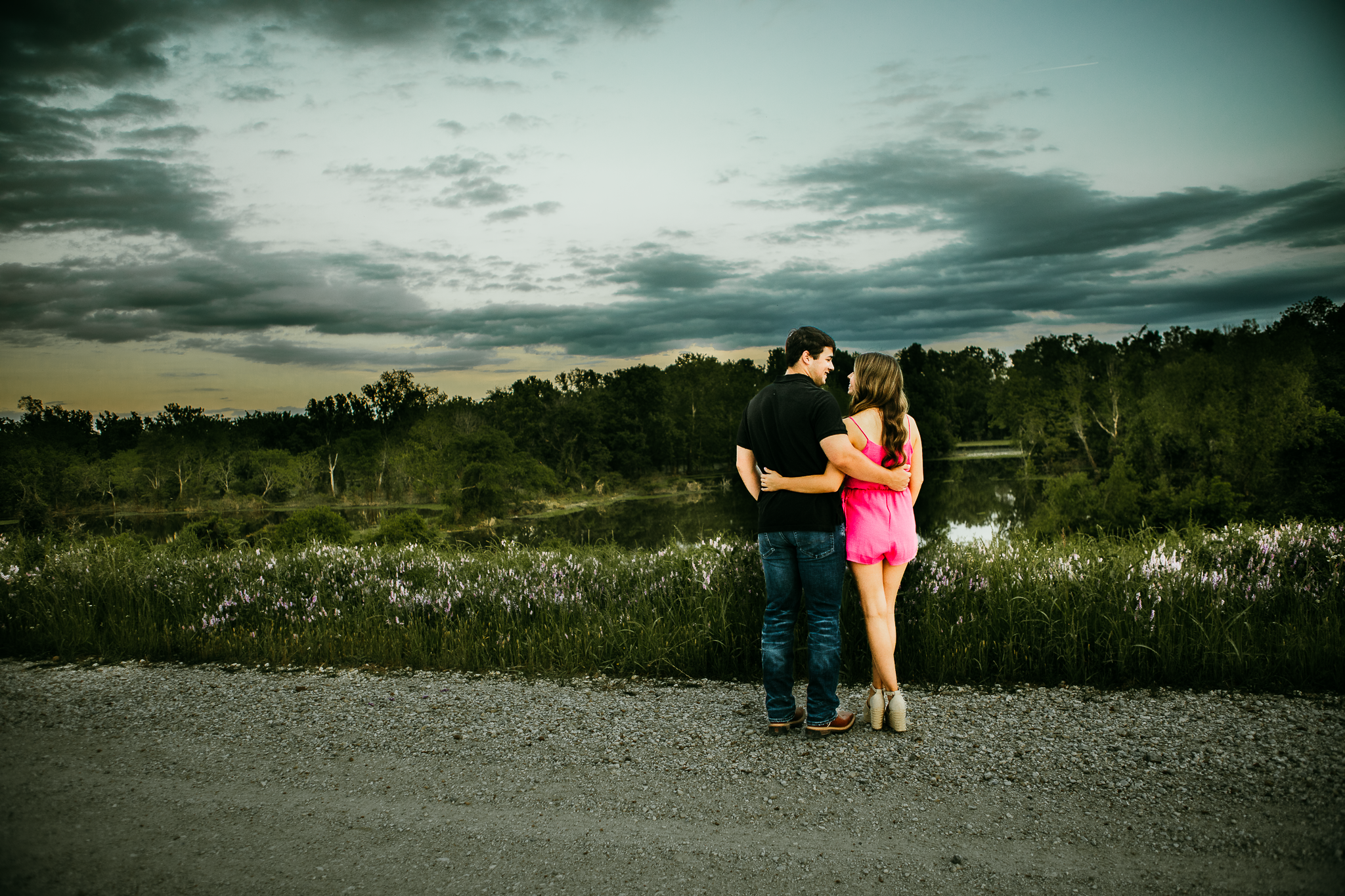 A young man and woman stand close together on a gravel path near a lake, with trees and cloudy sky in the background. The woman is wearing a pink dress and heels, and the man is in a black shirt and jeans. They are hugging and gazing at each other.