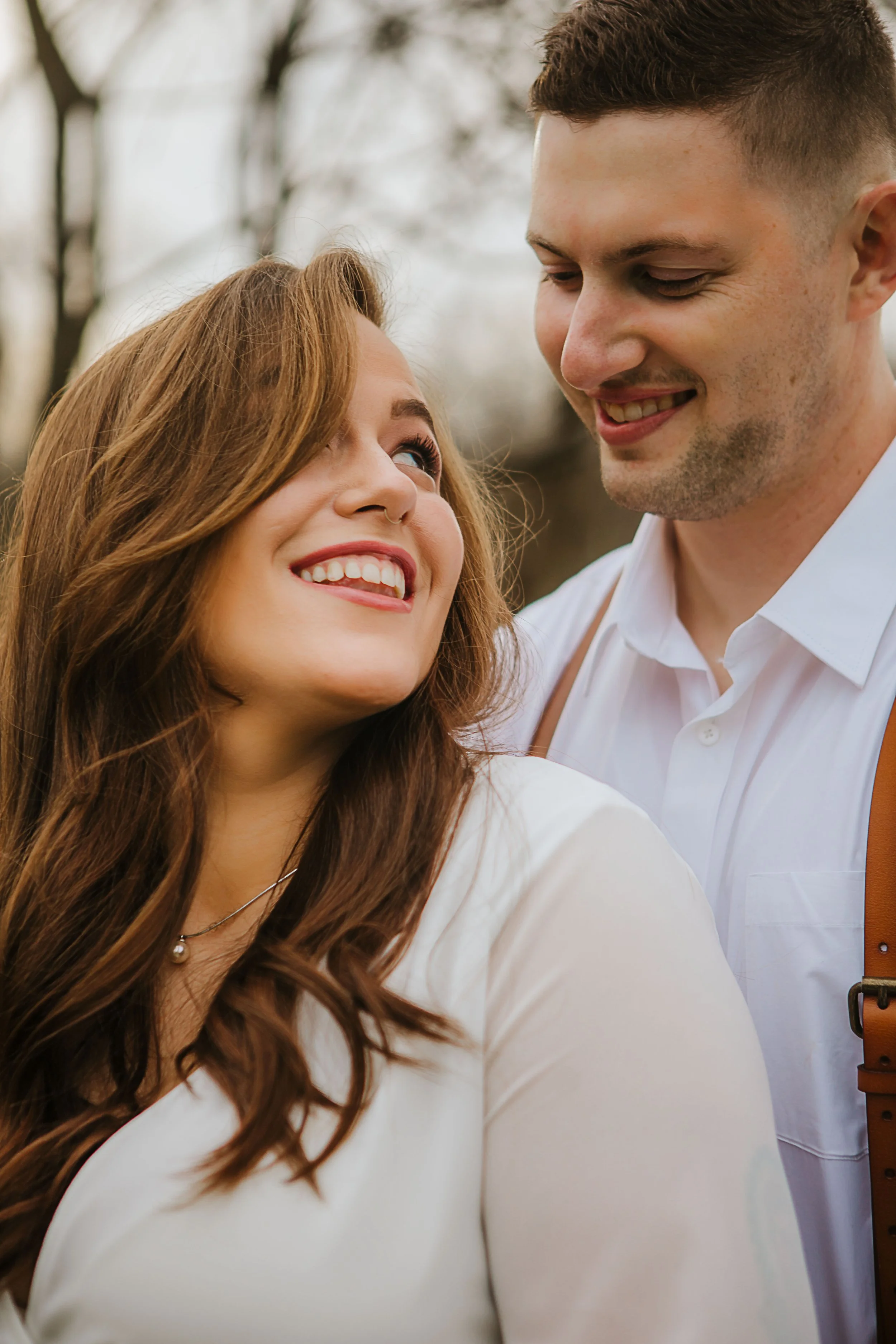 A woman with red hair and a man with short brown hair smiling and looking at each other outdoors.
