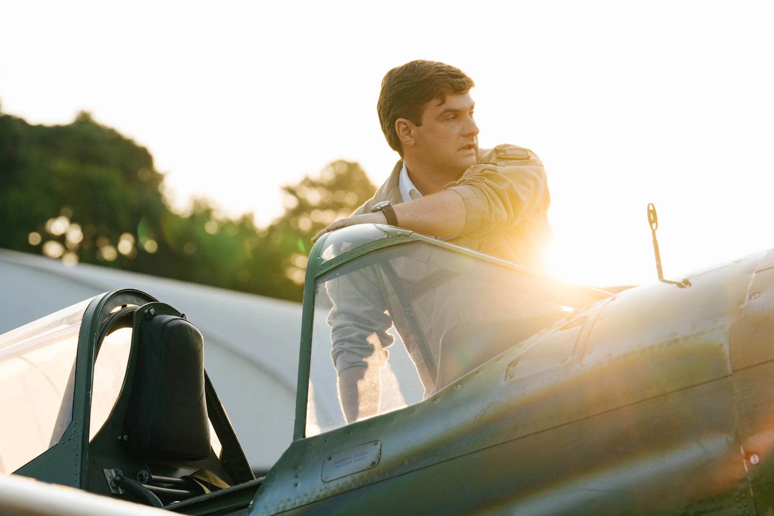 A man leaning on an open cockpit of a vintage aircraft with sunlight in the background.