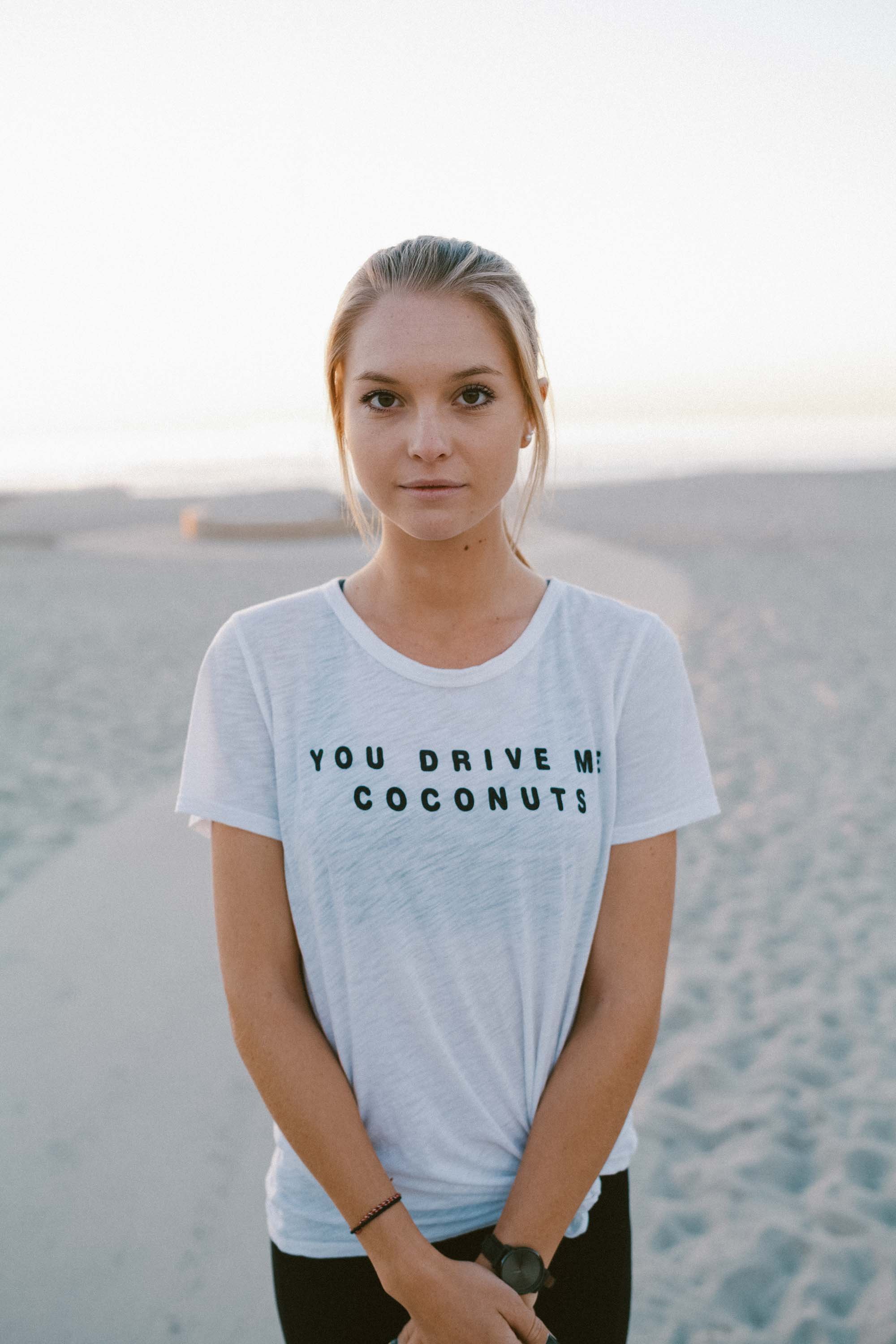 Person wearing a white T-shirt with text "You drive me coconuts" standing on a beach.