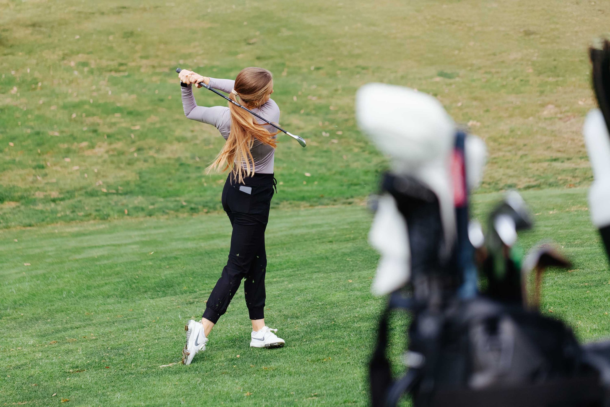 Woman in casual sportswear playing golf, swinging a club on a green golf course, with a golf bag in the foreground.