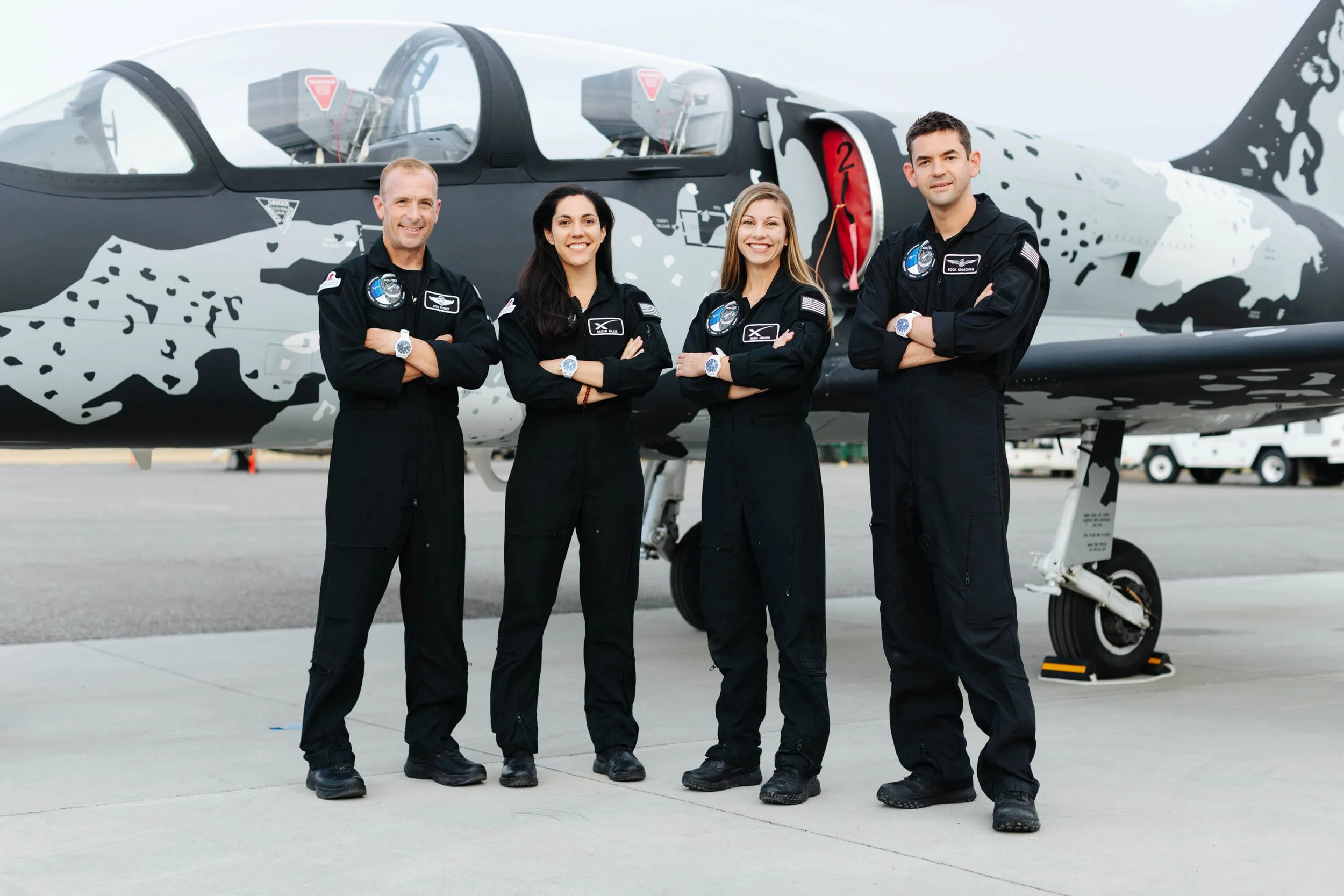 Four people in black flight suits standing in front of a sleek, camouflaged aircraft on a tarmac.