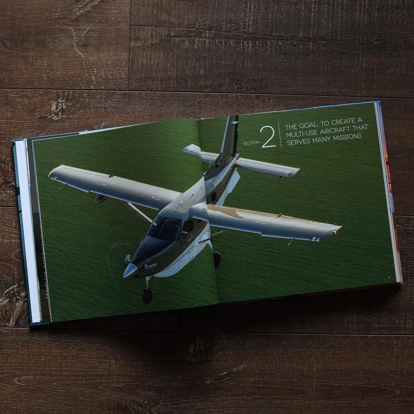 Open book on a wooden surface featuring an aerial view of an aircraft in flight over green terrain. The page contains the text "Section 2 The goal to create a multi-use aircraft that serves many missions."