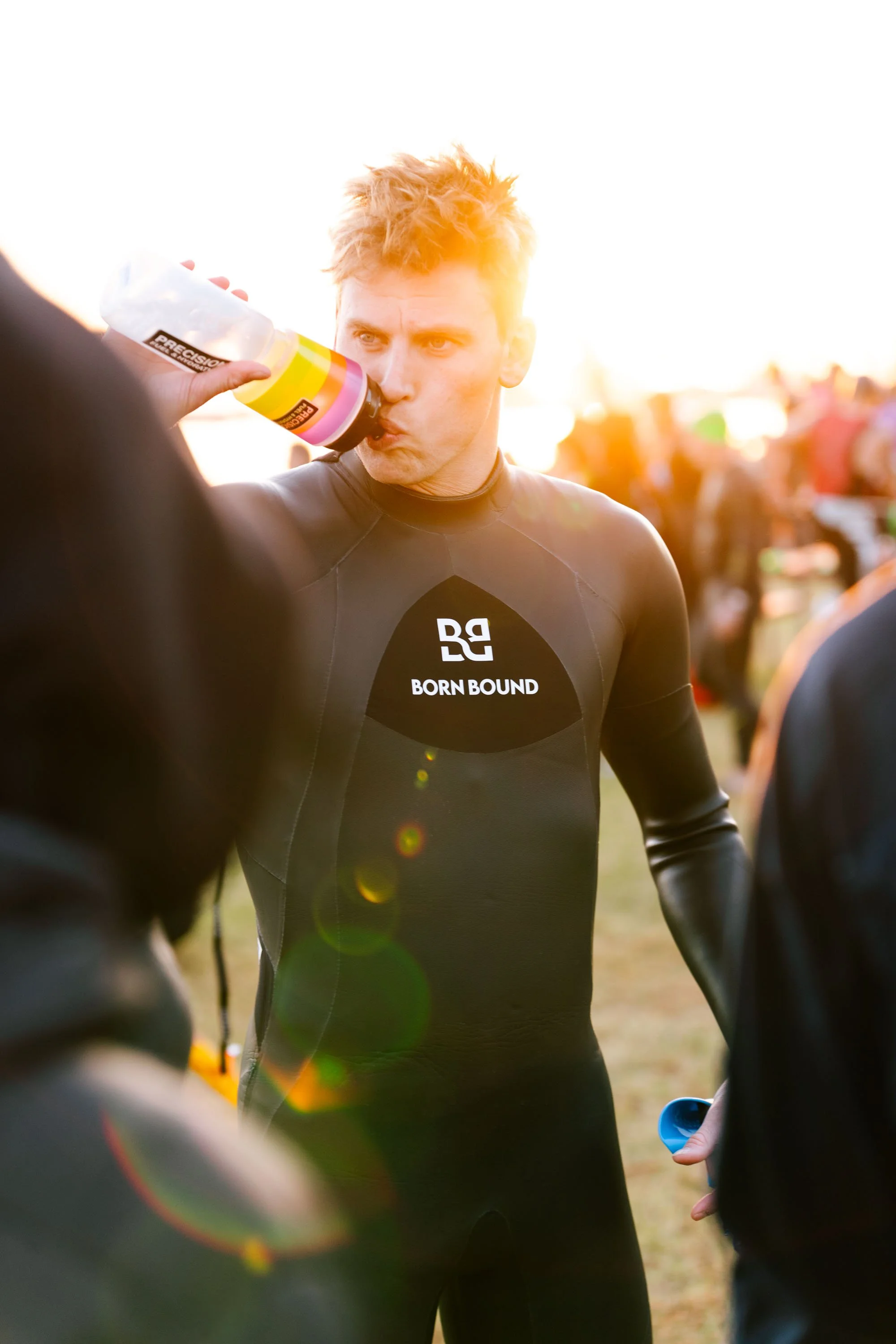 Person in a wetsuit drinking from a bottle outdoors during a sporting event.
