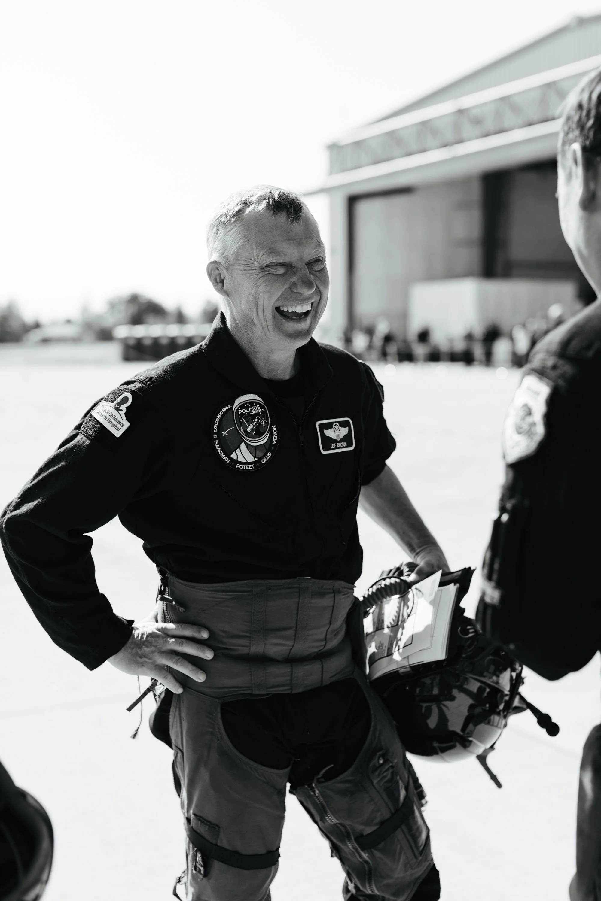 A person in a flight suit laughing and conversing with another individual at an airfield.