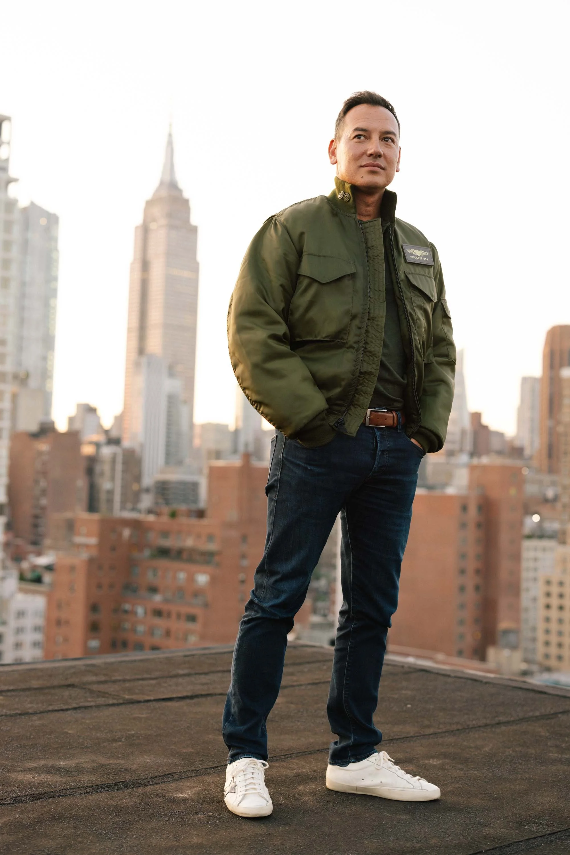 Man wearing a green jacket and jeans on a rooftop with a cityscape and Empire State Building in the background.