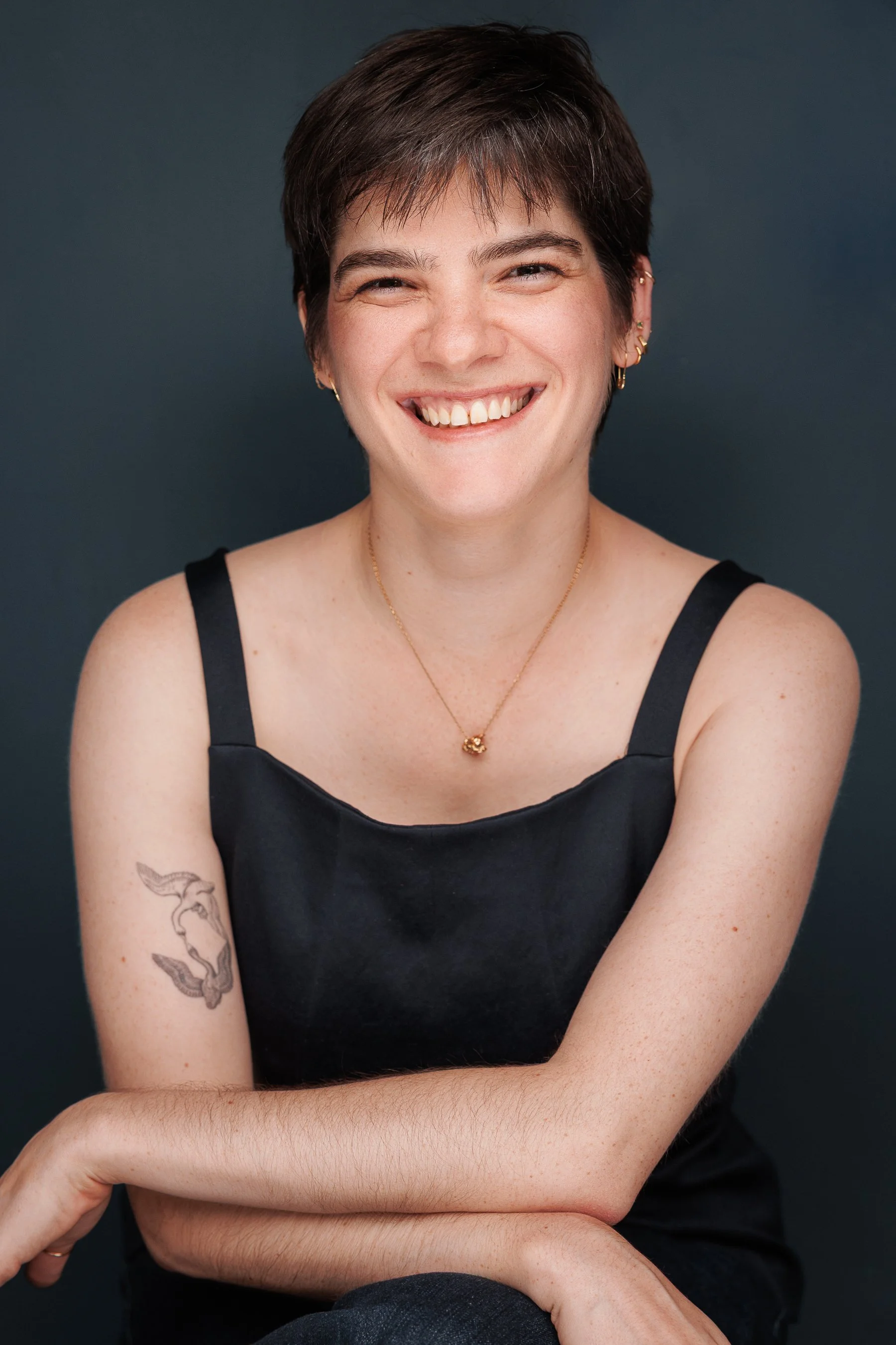 A young woman with short brown hair, smiling widely, wearing a black sleeveless top, gold jewelry, and tattoos on her arm, sitting against a dark background.