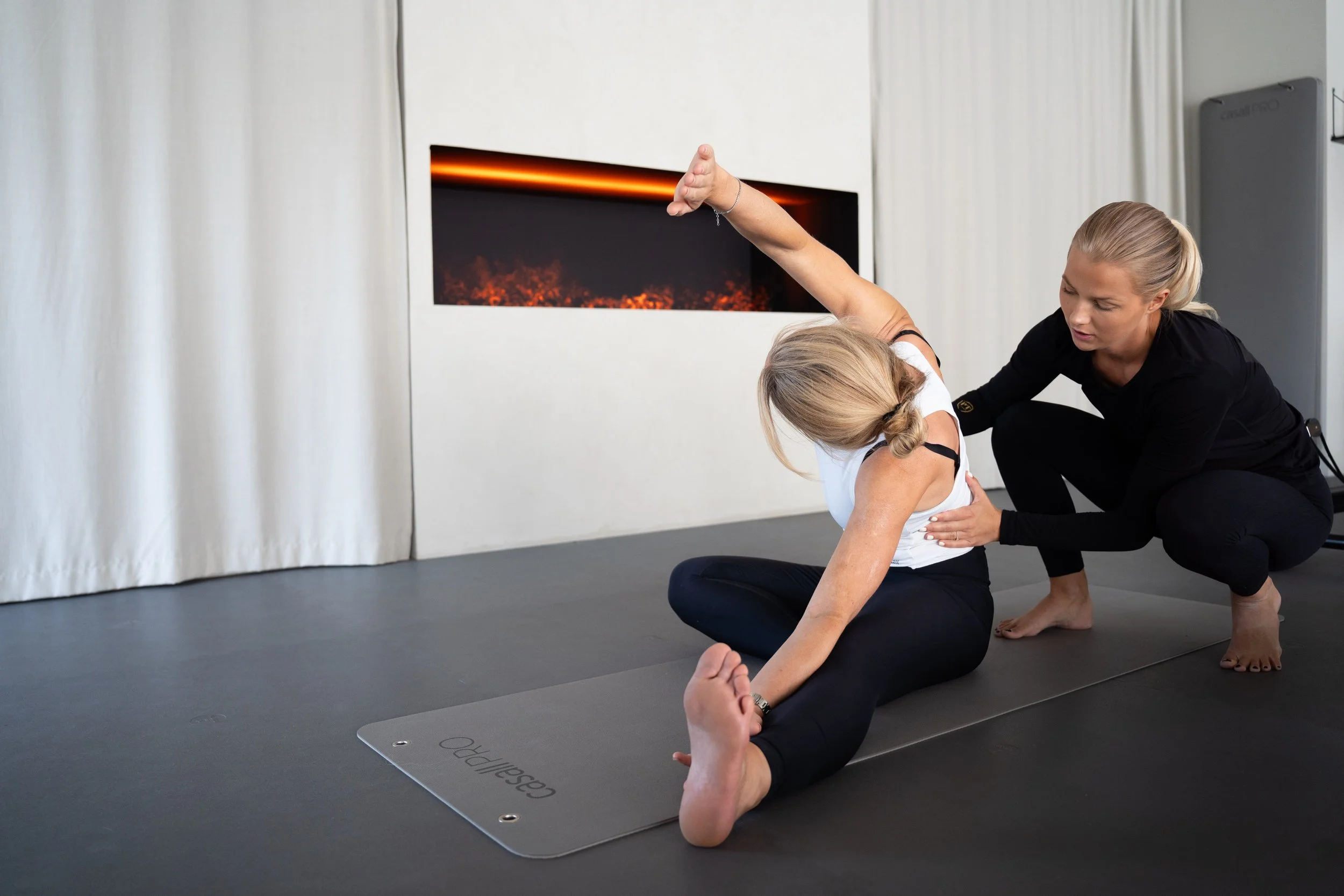 En kvinna hjälper en annan kvinna att göra en stretchingövning på en träningsmatta i ett rum, med en eldstad på väggen i bakgrunden.