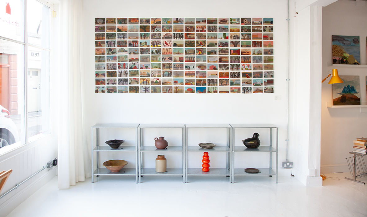 Gallery wall with a grid of small landscape paintings, metal shelving with ceramic and decorative objects, and a window with white curtains in a bright, minimalist art space.