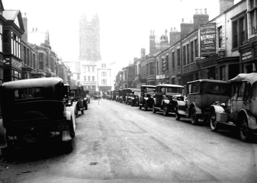 Black and white photograph of a city street with vintage cars lined up on both sides and buildings with signs, including a sign for plumbers and estate agents, in the background with a tower or cathedral spire visible.