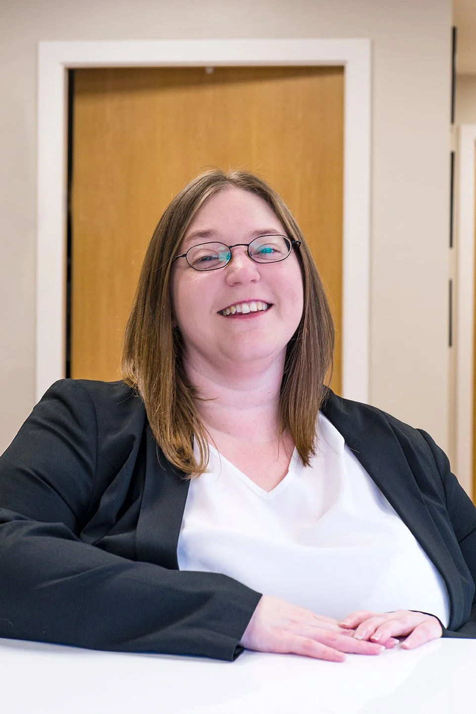 Smiling woman wearing glasses, black blazer, and white top sitting at a white table in an office setting.