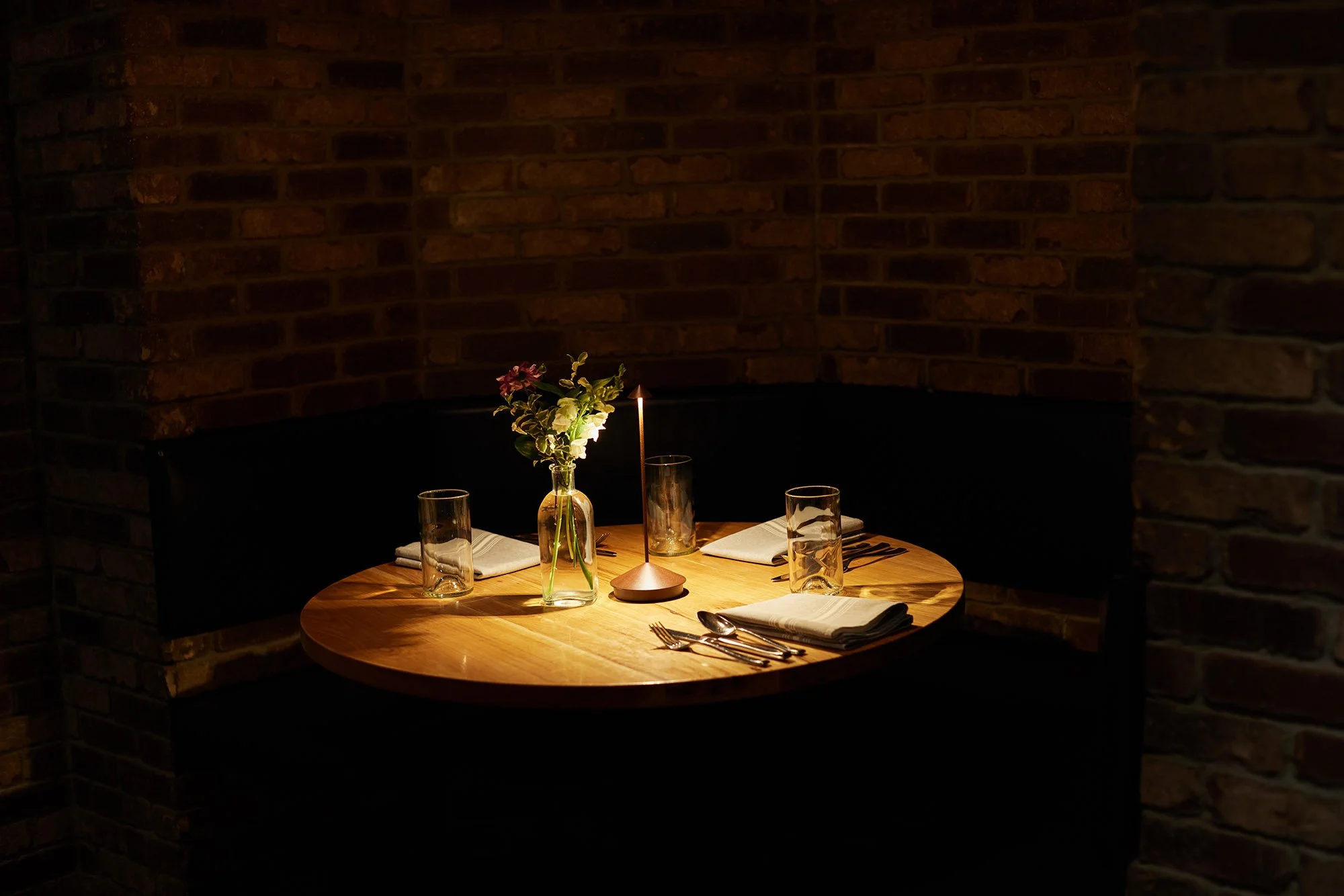 A dimly lit restaurant table set for four, with a small vase holding pink and white flowers, a candle, and white napkins with cutlery, against a brick wall background.
