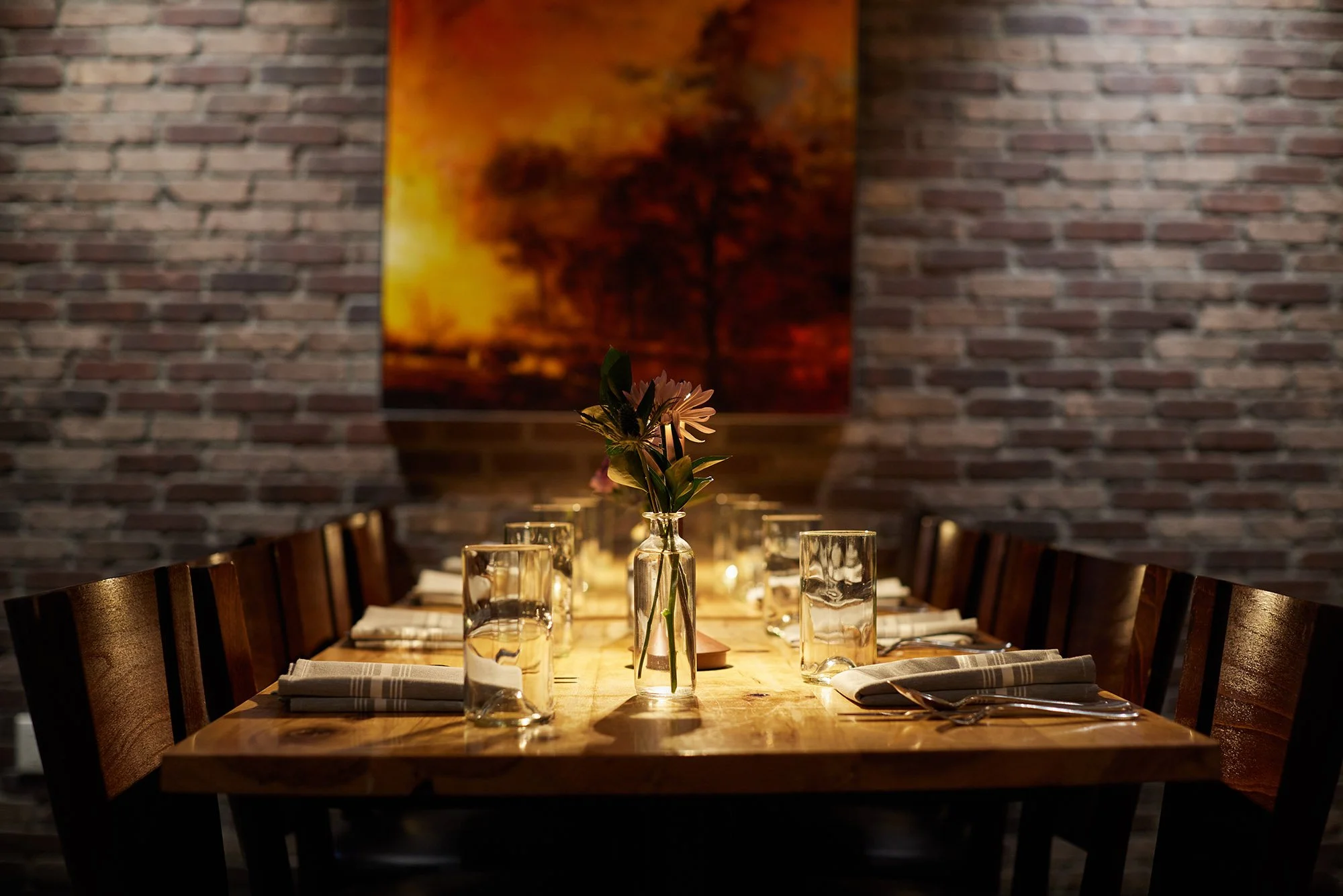 A dining table set with glasses, napkins, and a small vase with flowers, in a restaurant with a brick wall and abstract artwork in the background.