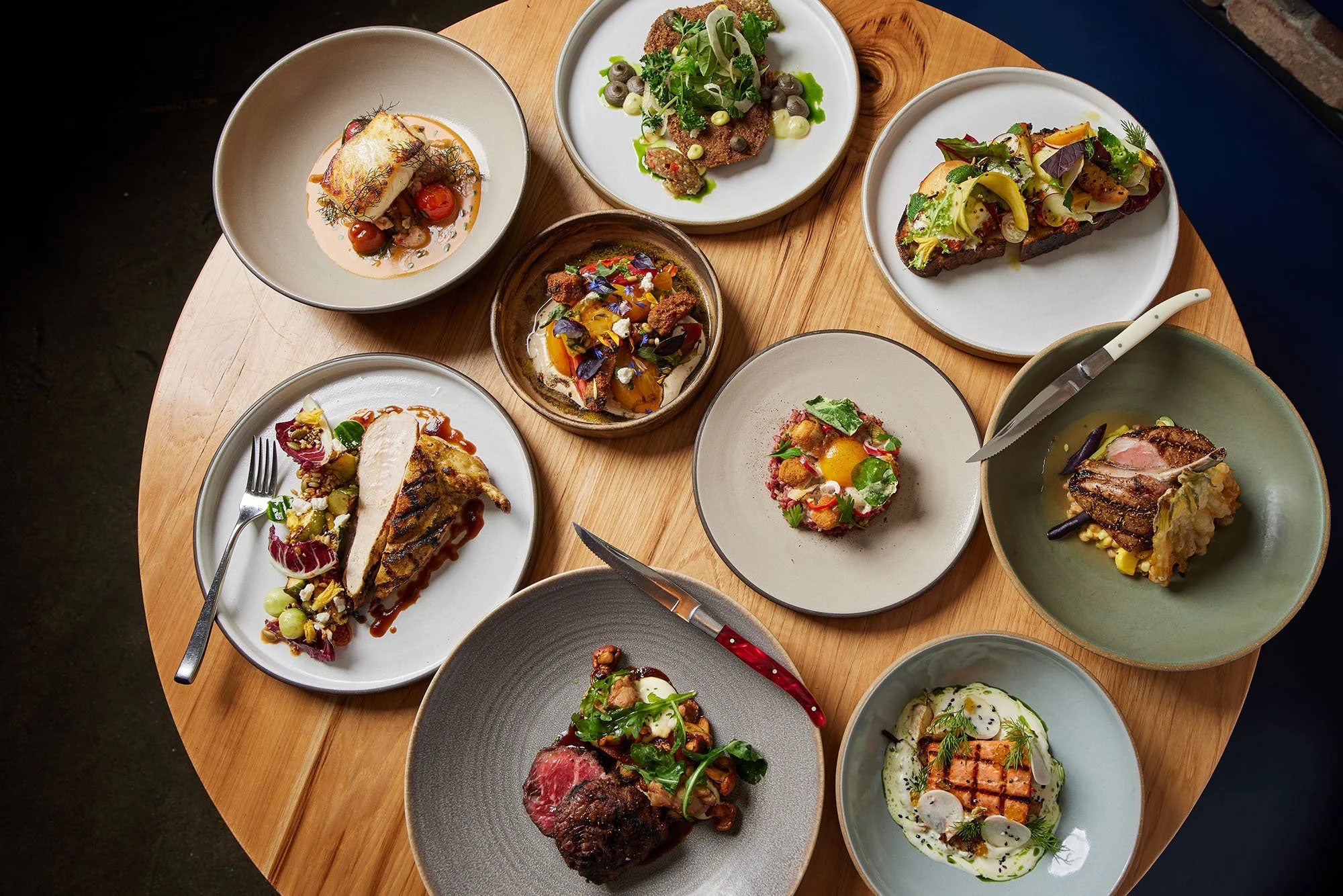 Assorted plated gourmet dishes on a wooden table, including roasted meat, salads, and other elegant appetizers.