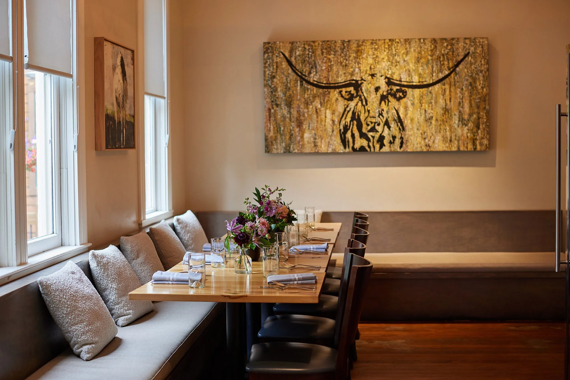 Interior of a restaurant with a long table set with glasses, napkins, and a flower arrangement, surrounded by chairs and a cushioned bench with pillows, against a wall decorated with artwork and windows with blinds.